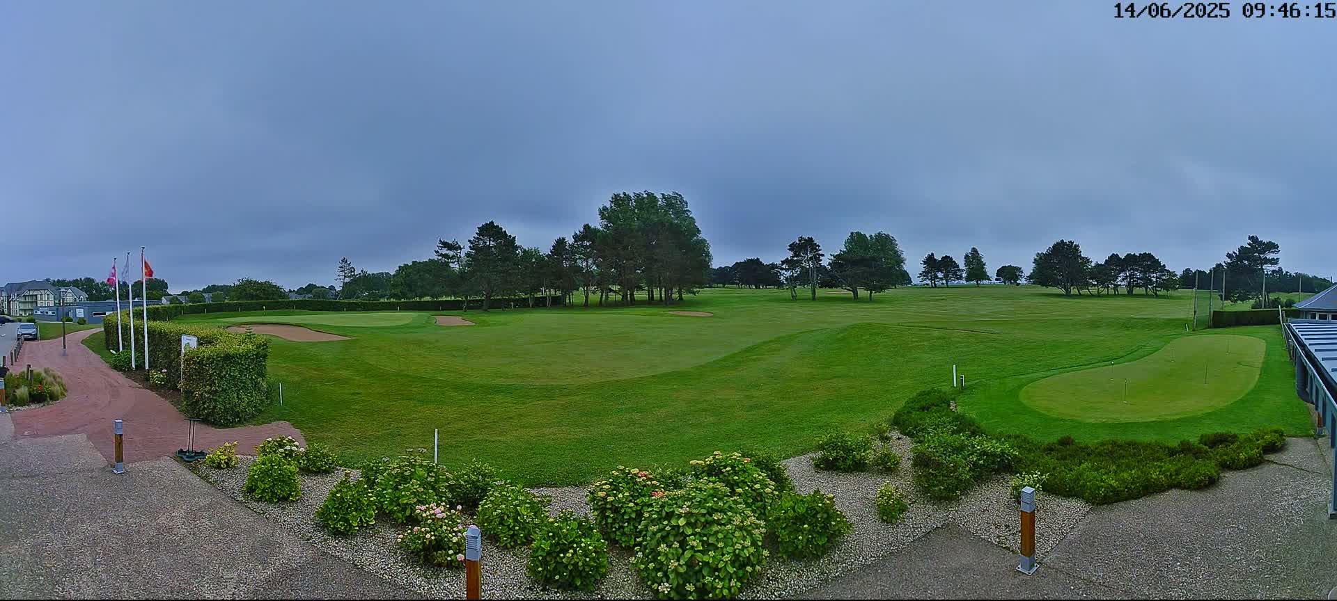 A golf course with green fairways and sand traps under an overcast sky is viewed from a paved area with landscaping.
