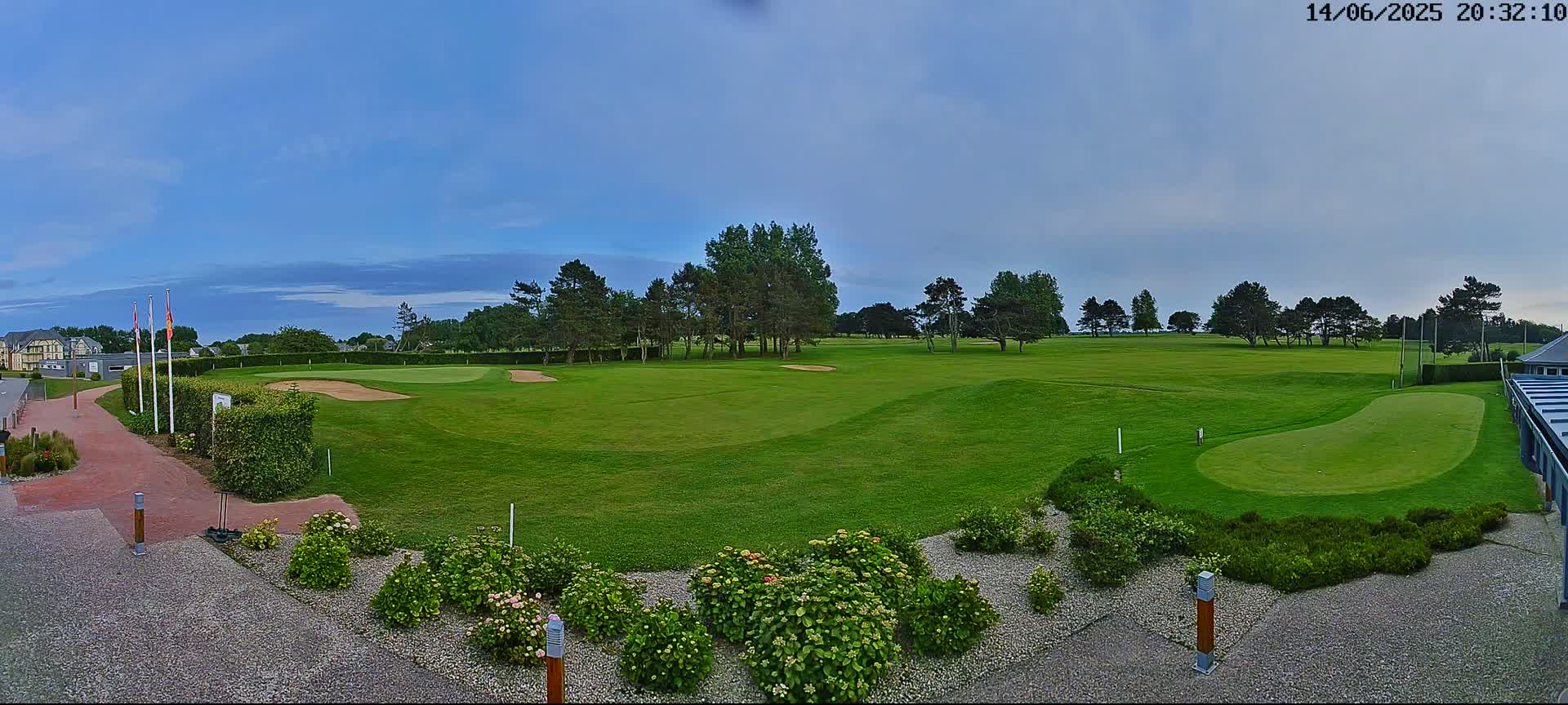 A panoramic view of a golf course under a mostly clear, twilight sky shows a putting green in the foreground, with a larger green and sand traps in the distance, surrounded by trees and landscaping.