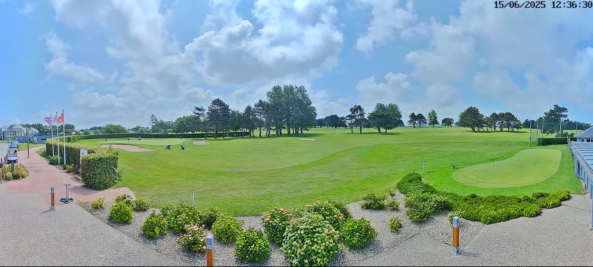 A sunny day on a golf course shows several golfers on the green, with a partly cloudy sky above.
