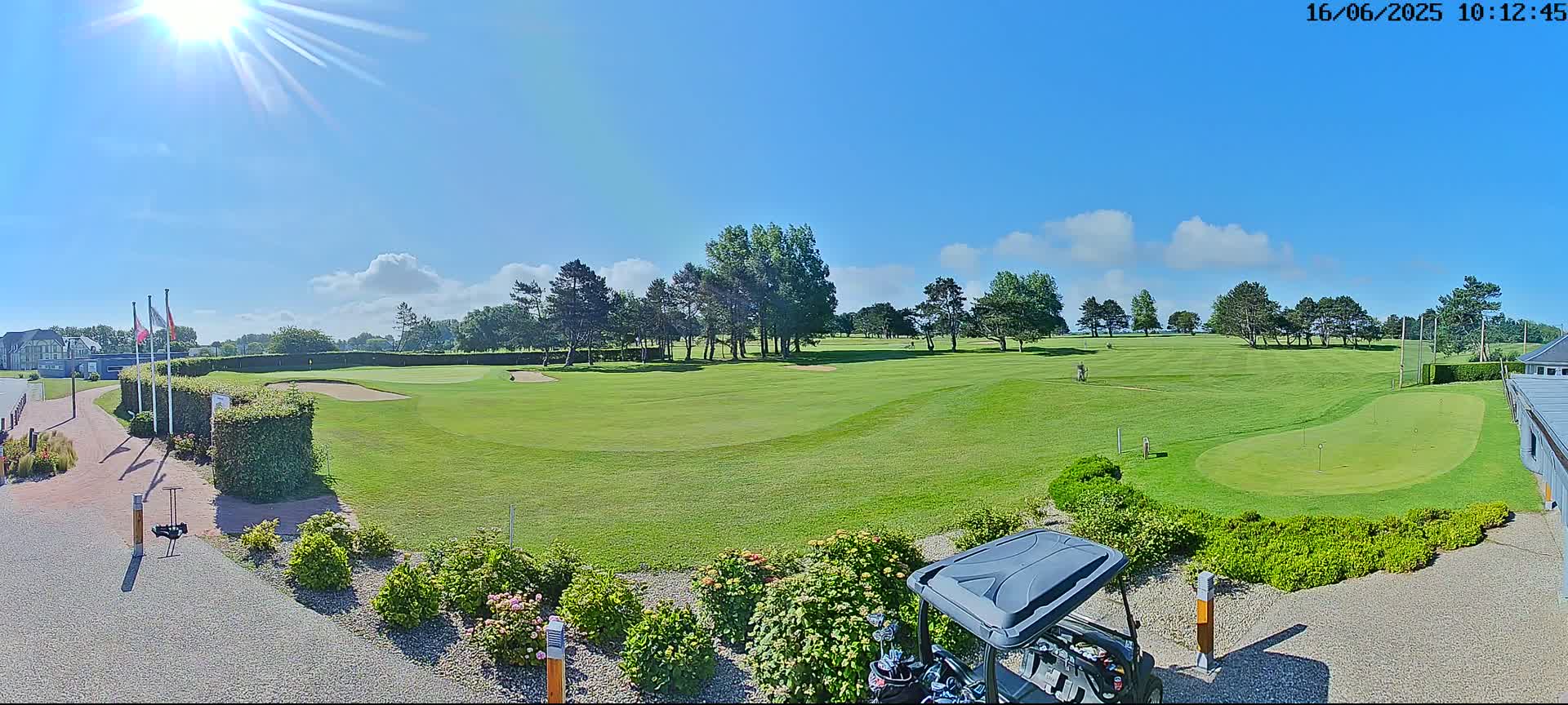 A sunny day on a golf course shows a putting green in the foreground, a fairway extending into the distance, and a golf cart parked near landscaping.