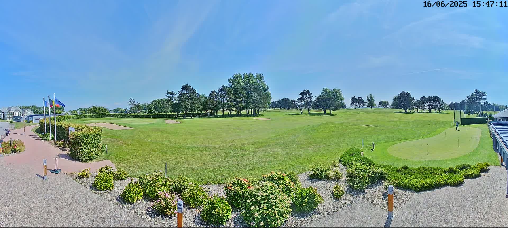 A sunny day on a golf course shows a putting green in the foreground and a fairway extending to a line of trees in the background.