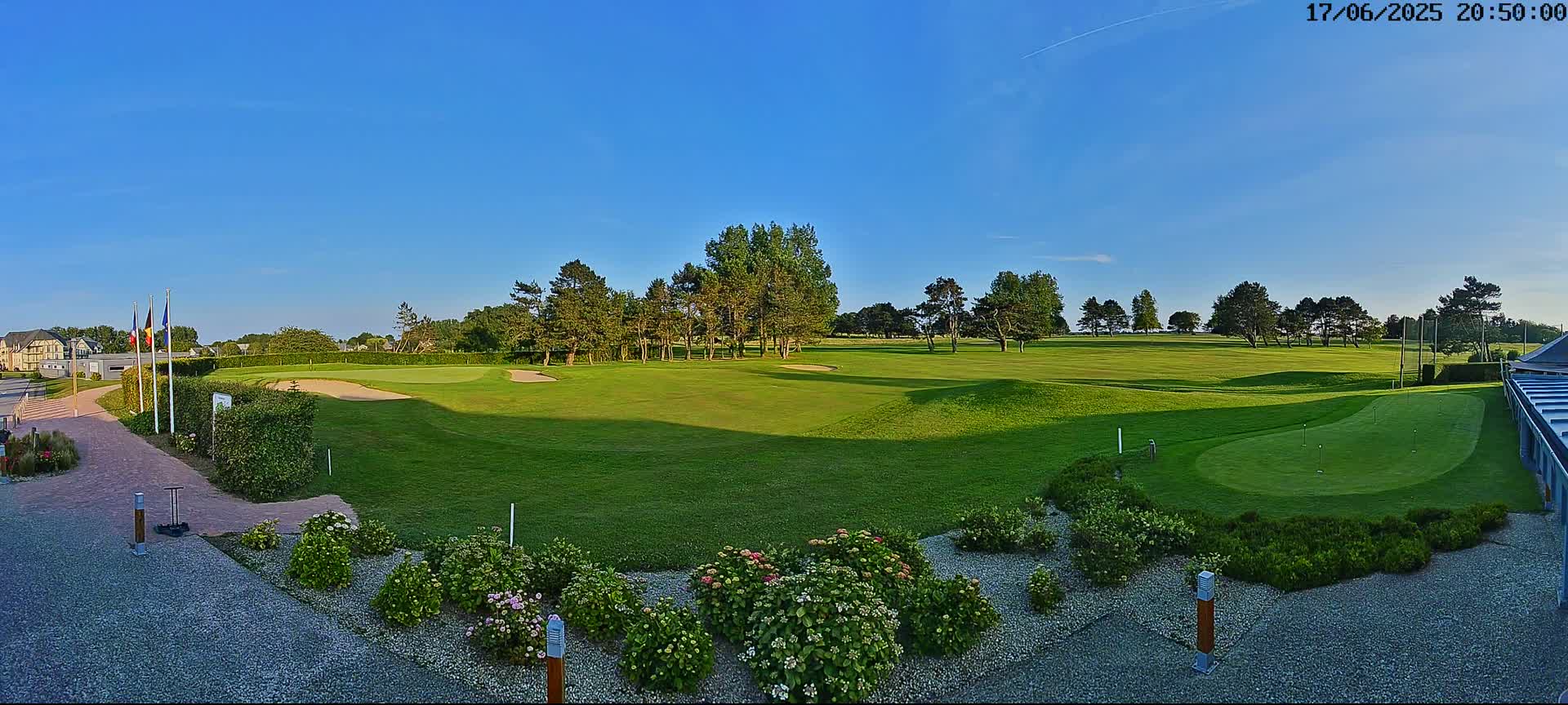 A sunny day reveals a verdant golf course with a putting green in the foreground, bordered by landscaping and a building.