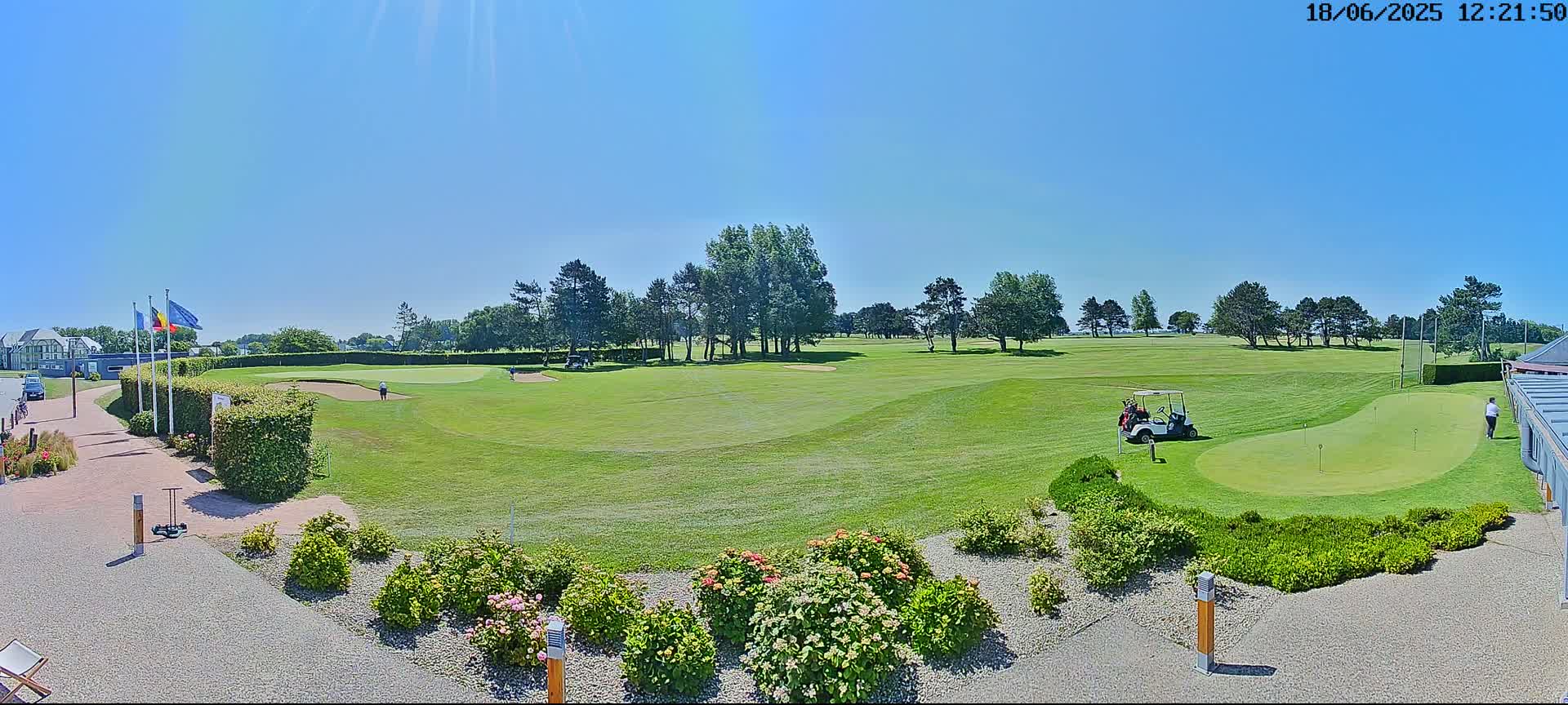 A sunny day on a golf course shows a putting green, golfers, and a golf cart near several trees and neatly landscaped grounds.