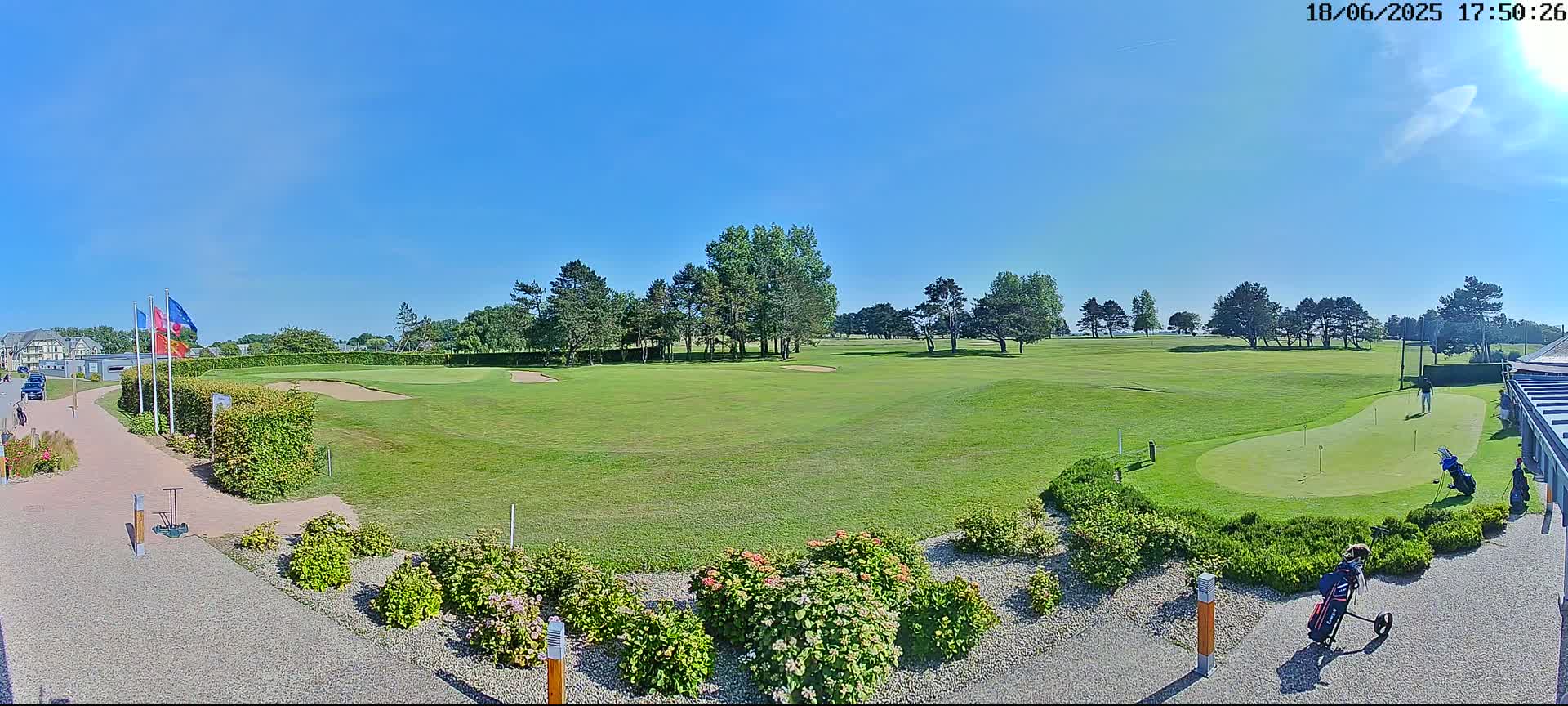 A sunny day at a golf course shows a putting green with golfers, landscaping, and flags.