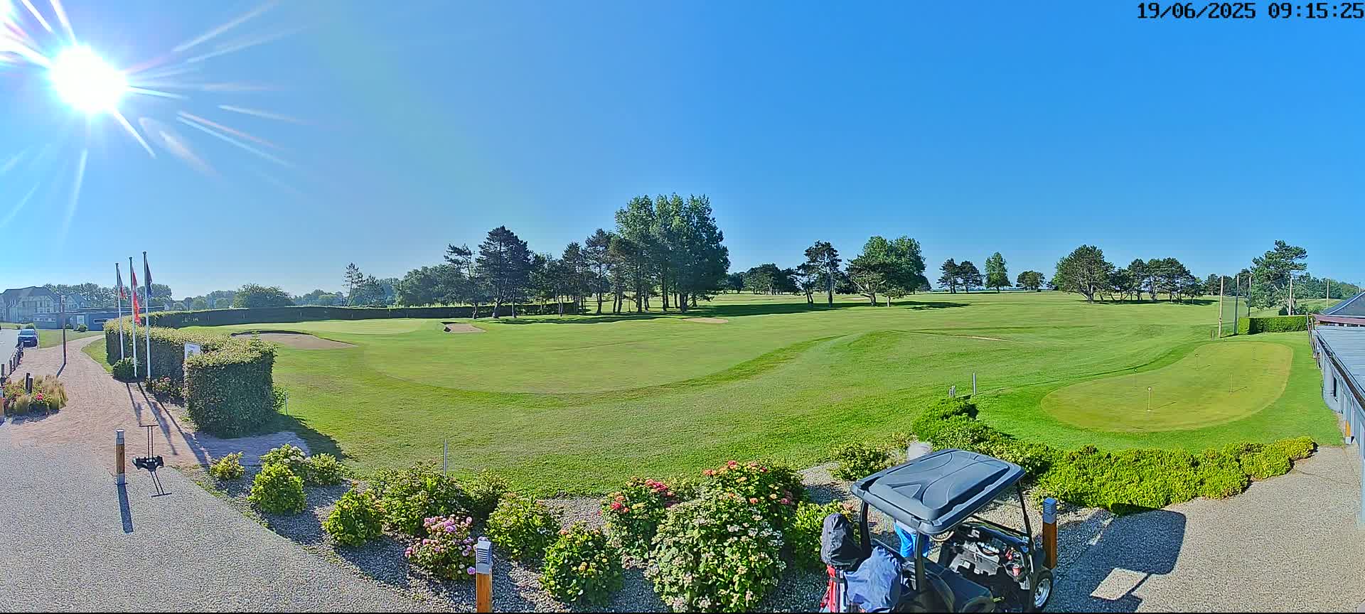 A sunny day on a green golf course, with a golf cart parked near a flowerbed in the foreground, and trees lining the fairways in the midground.