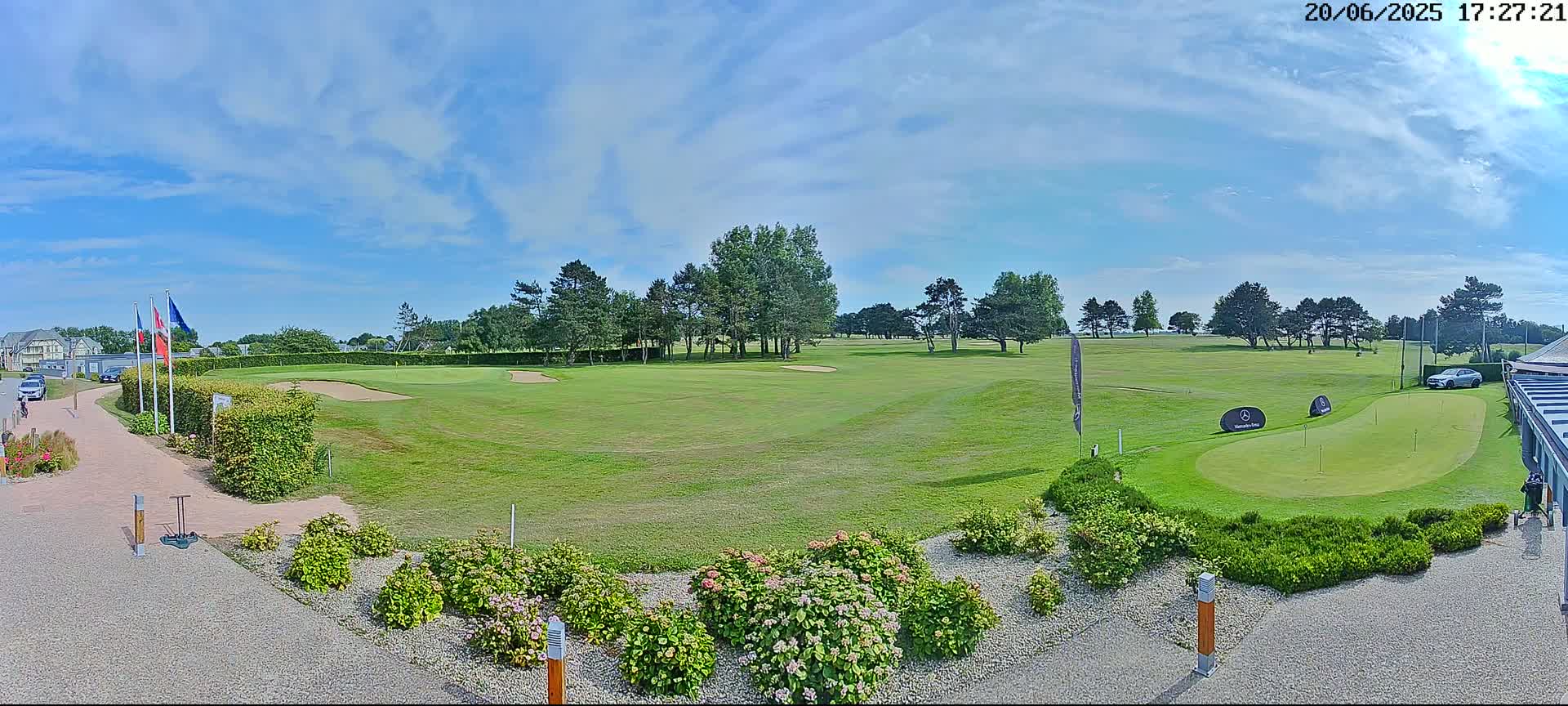 A sunny day on a golf course shows a putting green, a fairway, and several trees, with some shrubbery in the foreground.