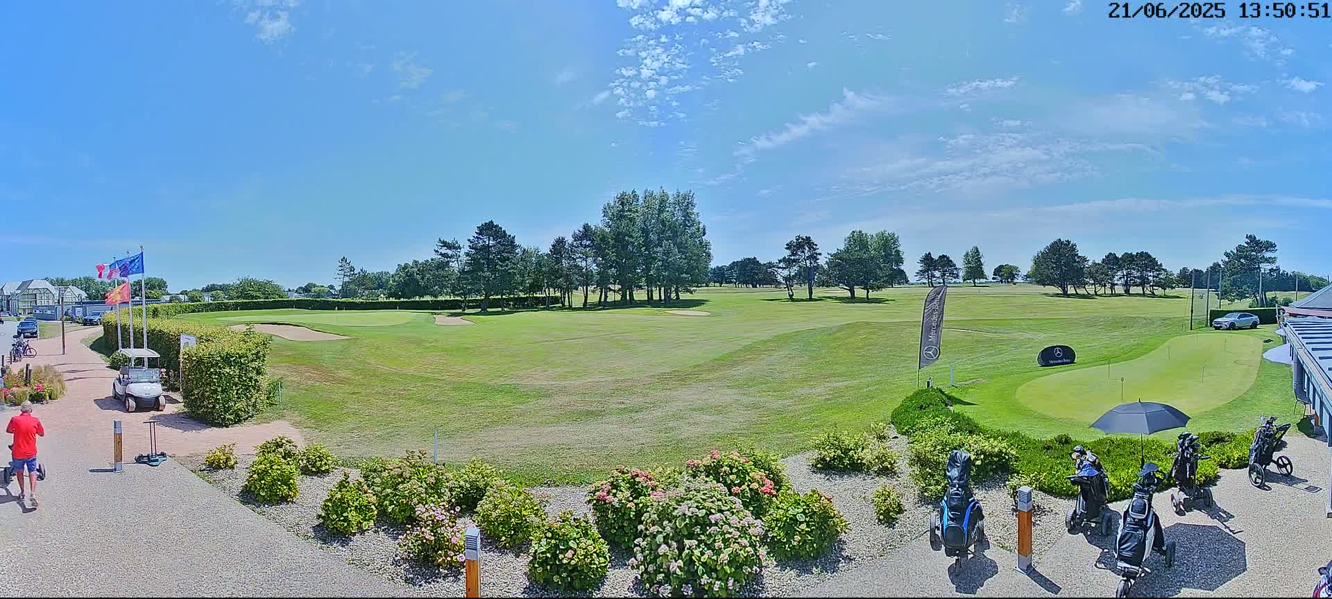 A sunny day on a golf course shows a putting green, several golf carts and bags, and a few people, all near a building.