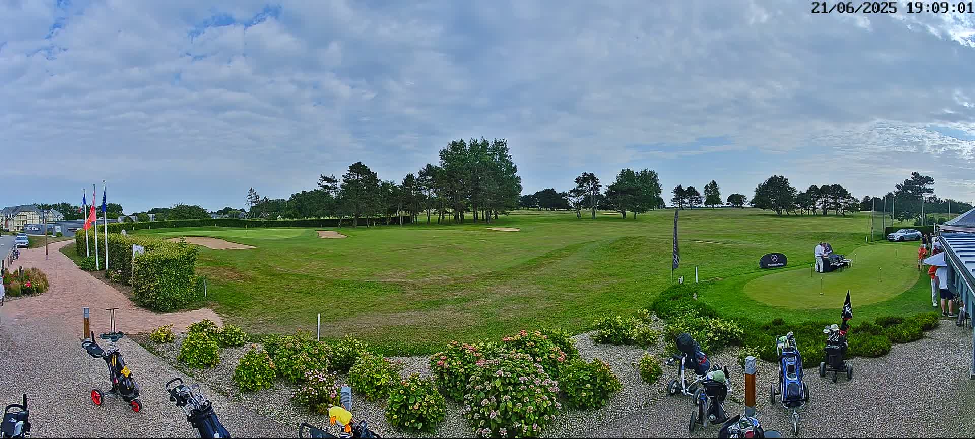 A partly cloudy day on a golf course shows a putting green with several people standing near a table, golf carts, and landscaping.