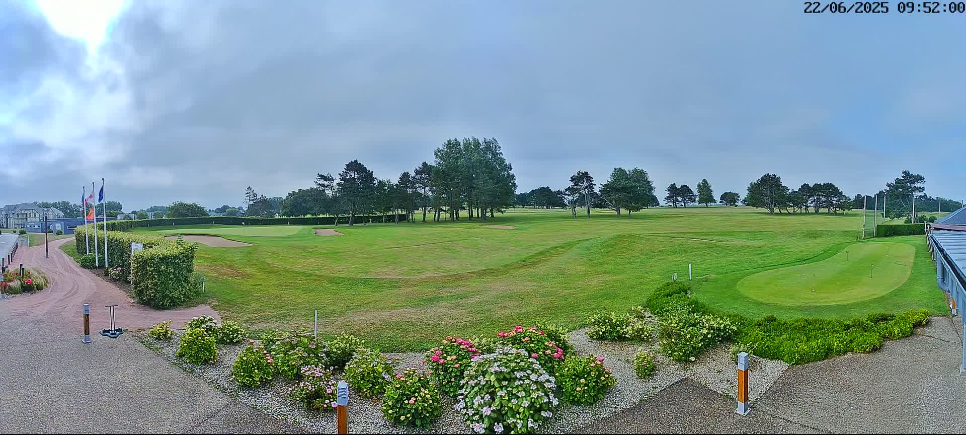 A green golf course with several sand traps and putting greens is shown under a mostly cloudy sky, bordered by a line of trees and colorful flowerbeds.