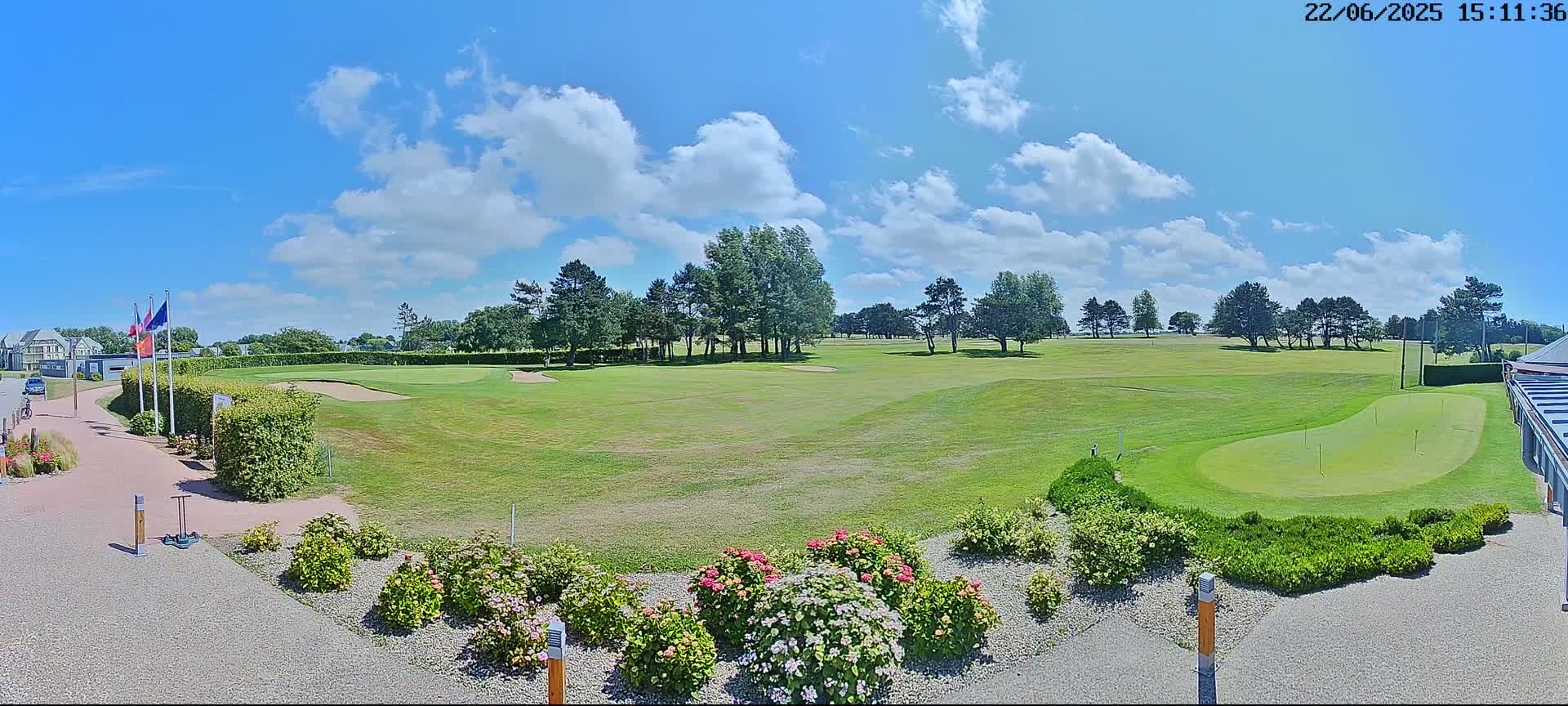 A sunny day with partly cloudy skies shows a golf course with green fairways, sand traps, and a putting green, bordered by trees and flowering bushes.