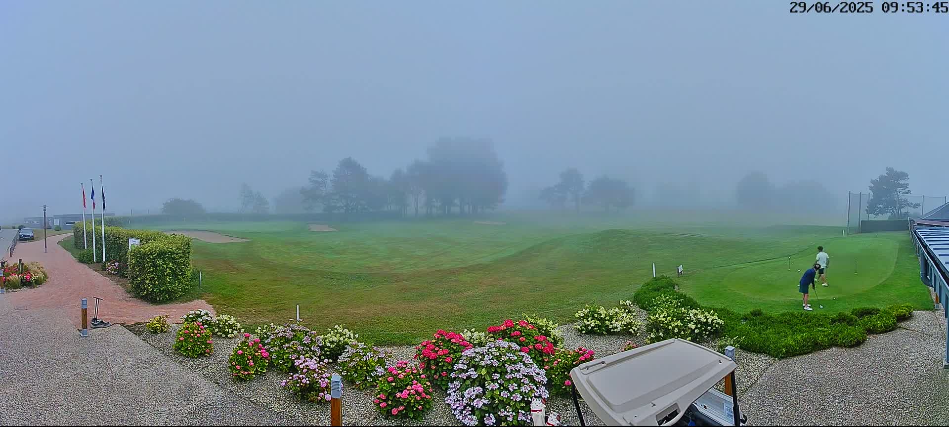 A foggy golf course with two people playing on a putting green, bordered by colorful flowers and a golf cart.