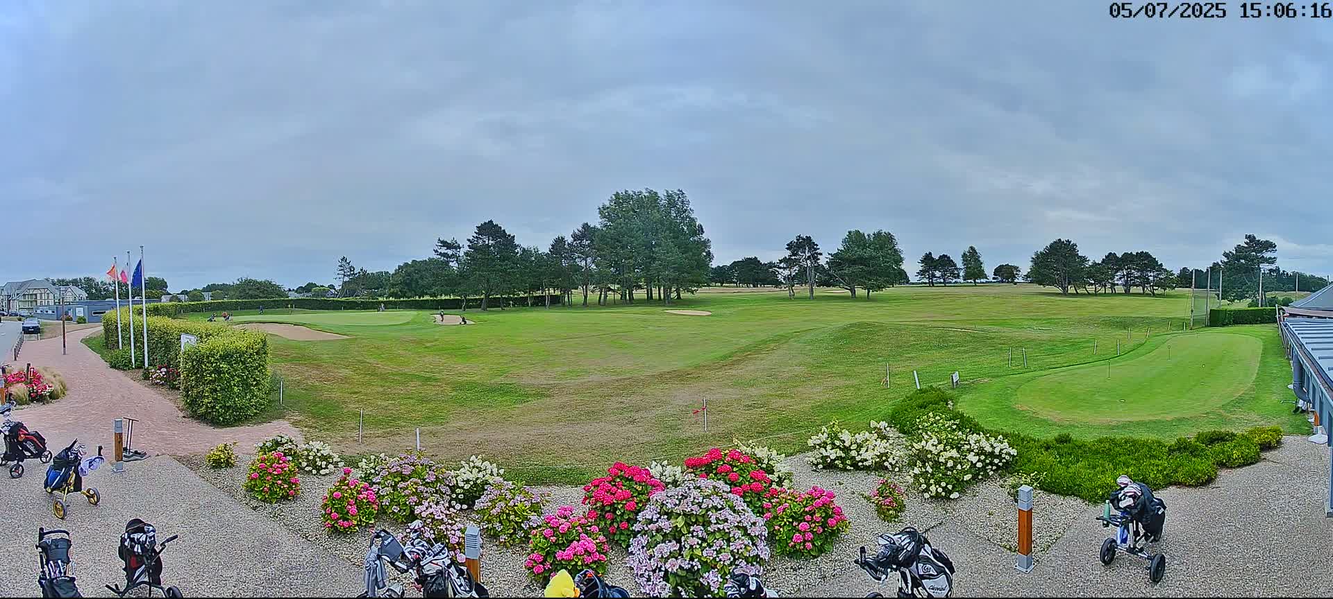 A golf course with several golfers visible in the distance, under a cloudy sky, features a putting green in the foreground bordered by colorful flowers and golf carts near a building.