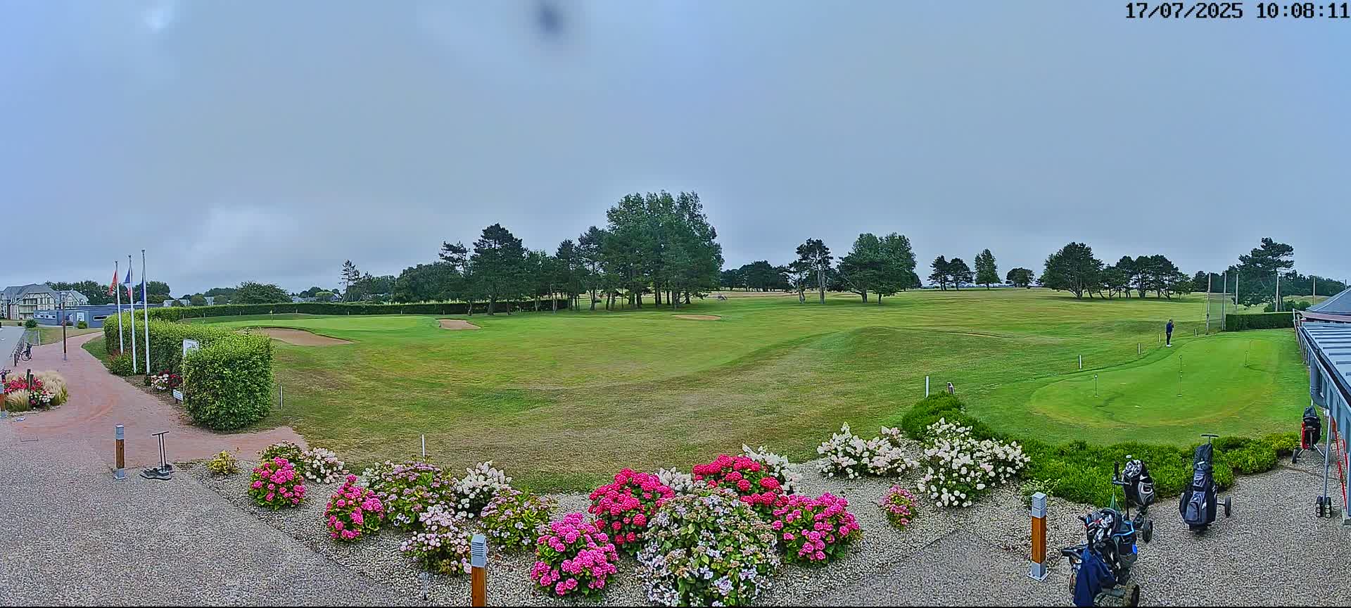A golf course with a putting green in the foreground, surrounded by flowers and trees under a cloudy sky.