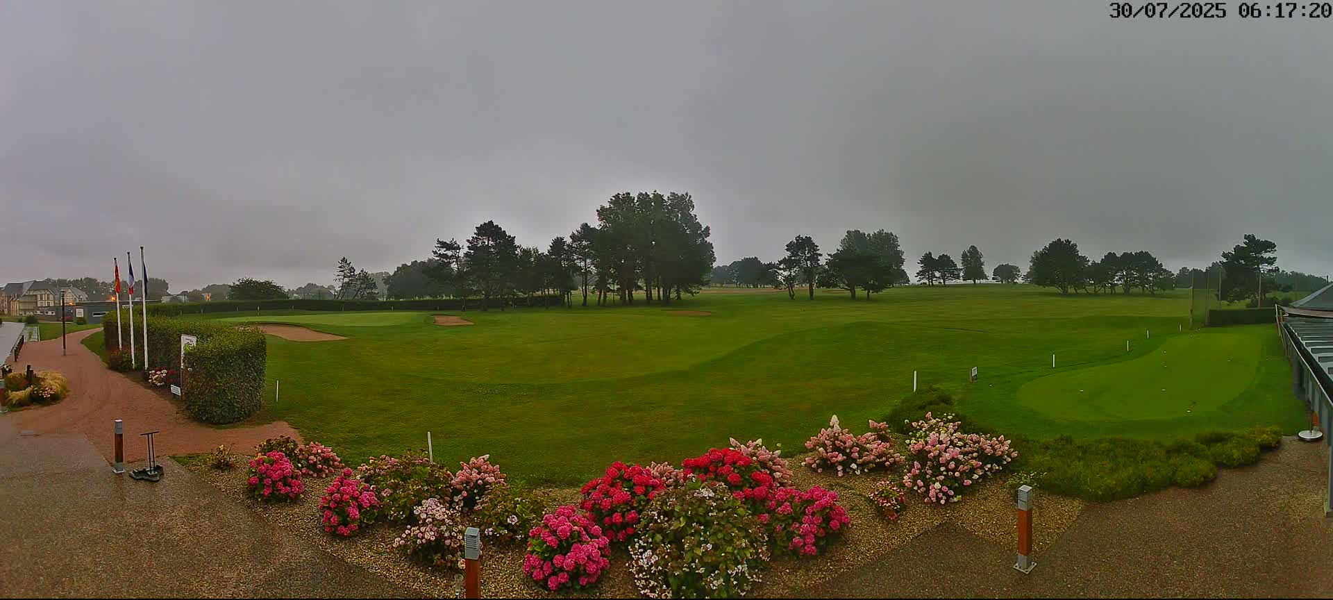 A golf course with a putting green in the foreground, surrounded by flowers and trees under a cloudy sky.