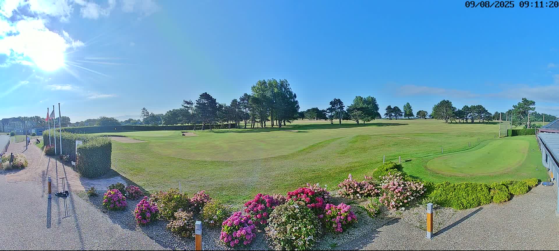 A sunny day reveals a golf course with green fairways, trees, and vibrant pink flowers in the foreground.
