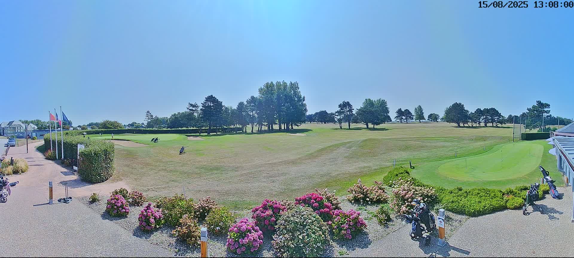 A sunny day reveals a golf course with green fairways, trees, and vibrant pink flowers in the foreground.