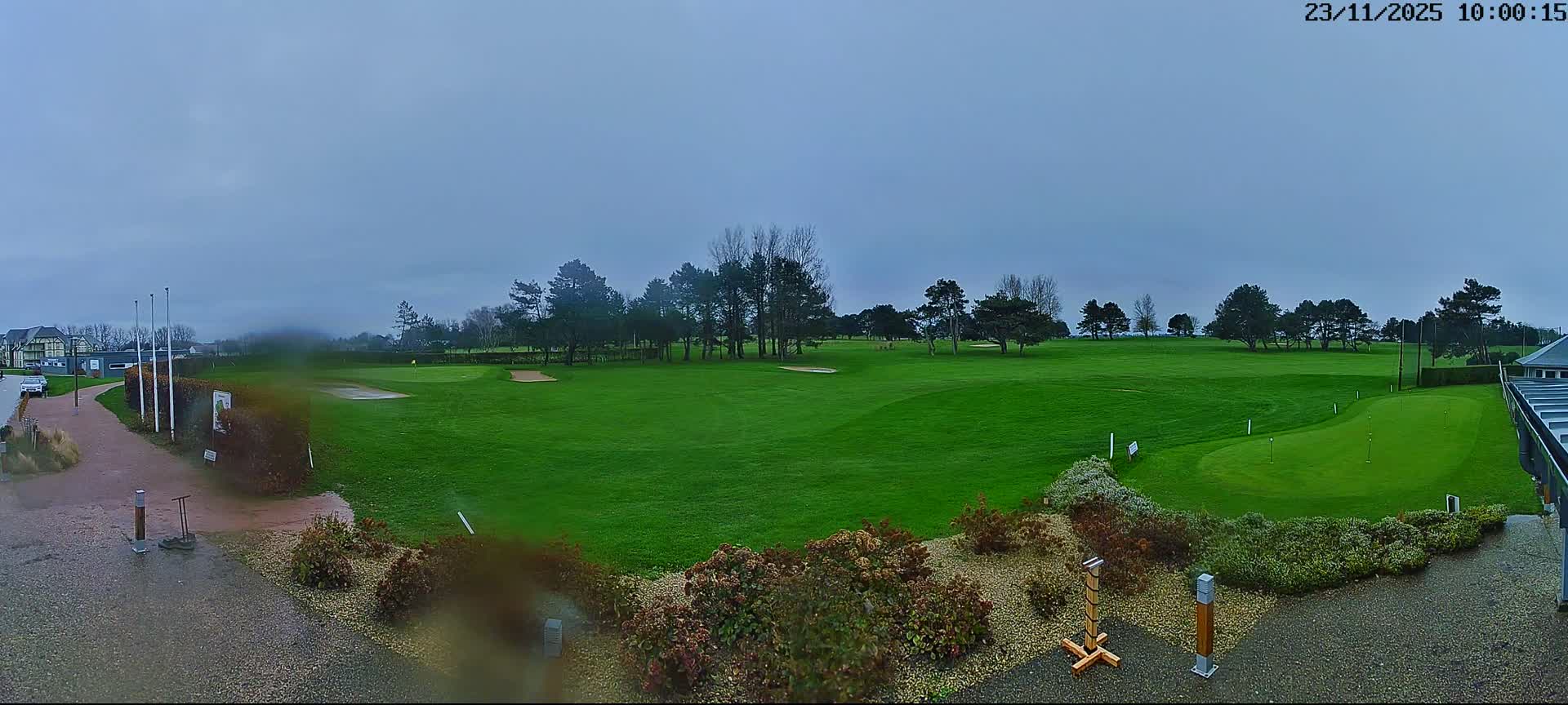 A wide view of a vibrant green golf course with bunkers and numerous trees unfolds under a dreary, overcast sky, with visible rain wetting the foreground path and ground.