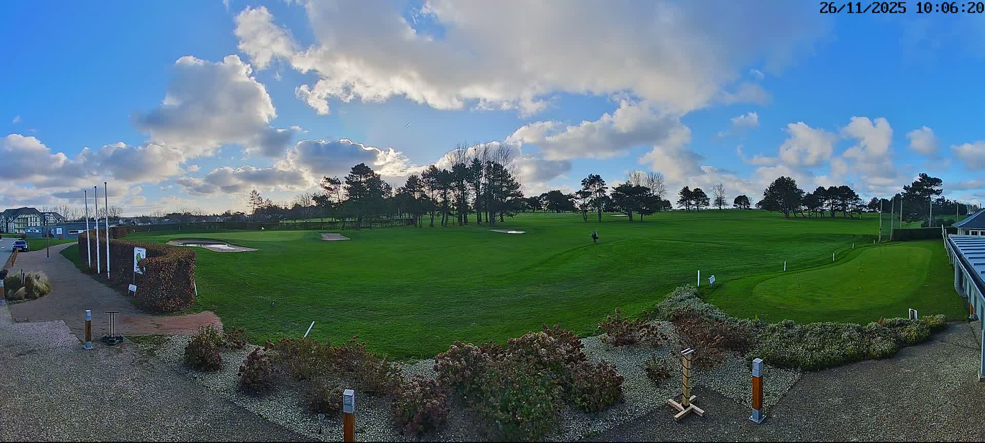 A wide view shows a vibrant green golf course with scattered trees, bunkers, and a distant golfer, all under a bright blue sky dotted with fluffy white clouds.