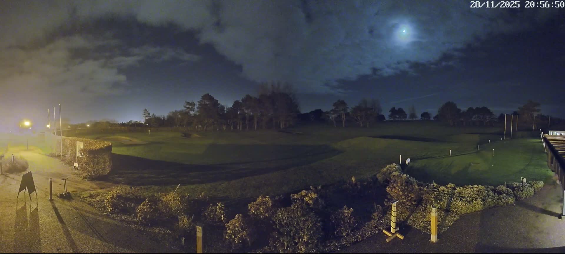A golf course is seen at night under a partly cloudy sky with a brilliant moon, while artificial lights illuminate paths and parts of the course.