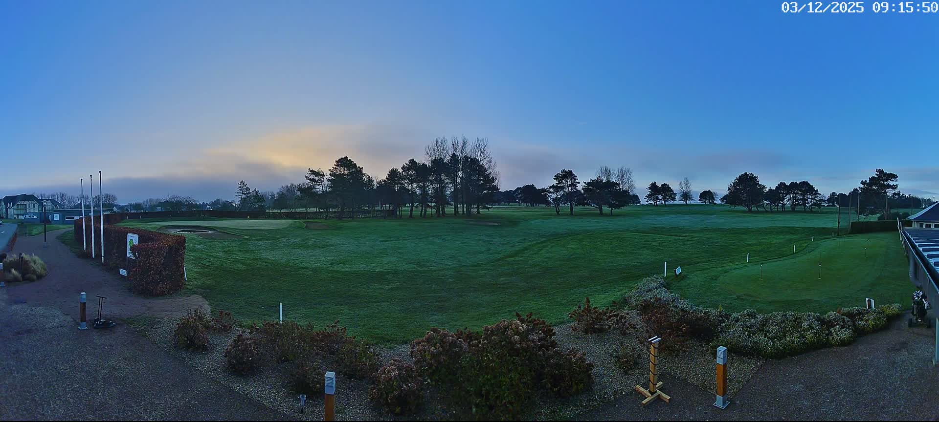 A panoramic view of a golf course features dewy green fairways, scattered trees, and distant buildings under a partly cloudy sky with patches of low-lying fog, suggesting a cool morning.