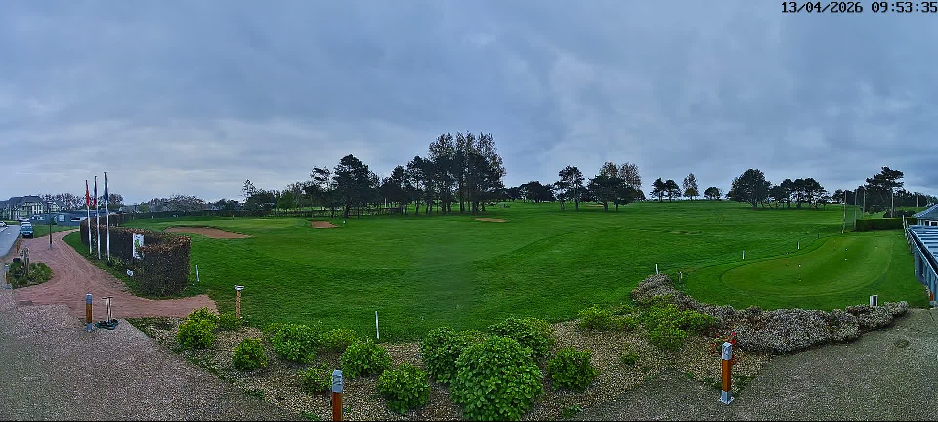 A panoramic view of a golf course features dewy green fairways, scattered trees, and distant buildings under a partly cloudy sky with patches of low-lying fog, suggesting a cool morning.