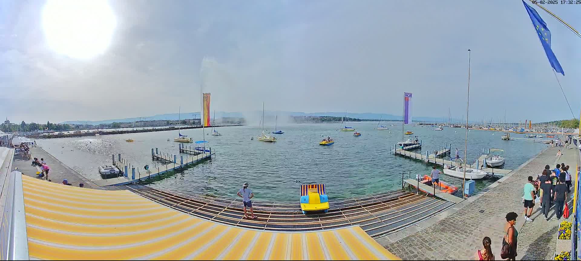 A sunny day at a harbor with several boats and people on the docks, and a large fountain spraying water in the background.