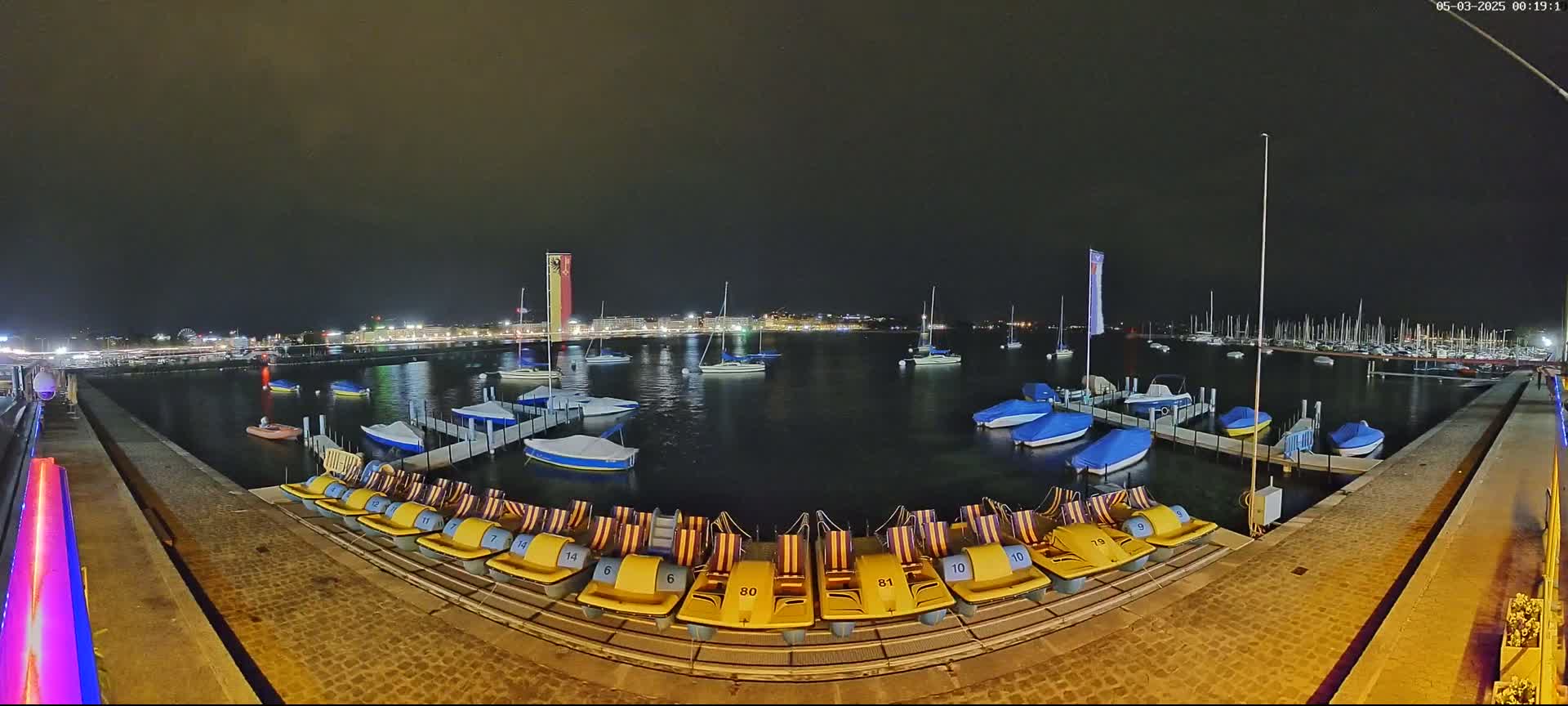 At night, a calm body of water has numerous docked boats, some covered, and a row of yellow paddle boats lined up along a stone dock with a city skyline in the background.