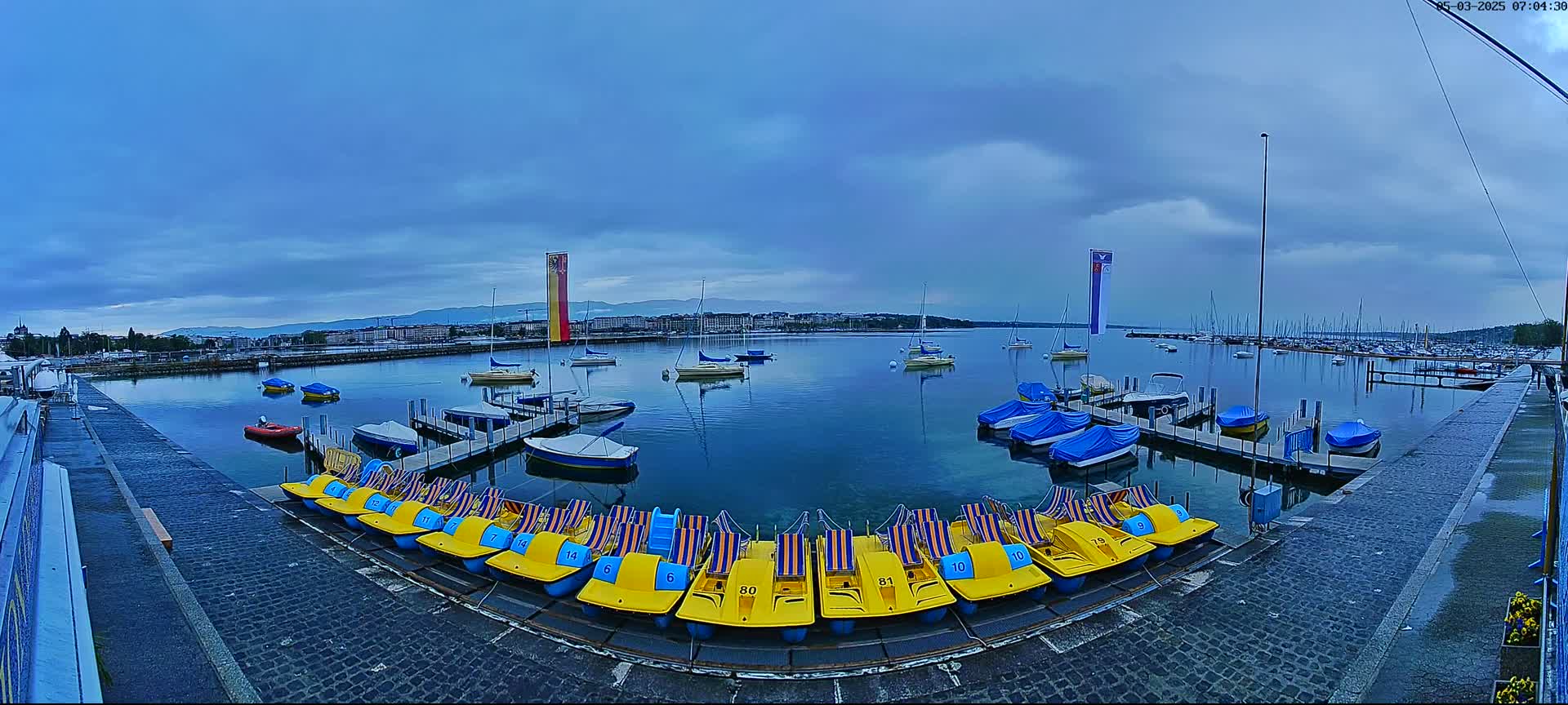 A calm lake with numerous boats moored at docks, under a cloudy, overcast sky, with a row of yellow paddle boats lined up along a stone pathway.