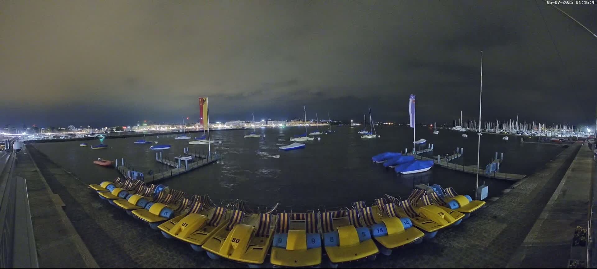 A nighttime view of a harbor with numerous boats and pedal boats docked, under a dark, cloudy sky.