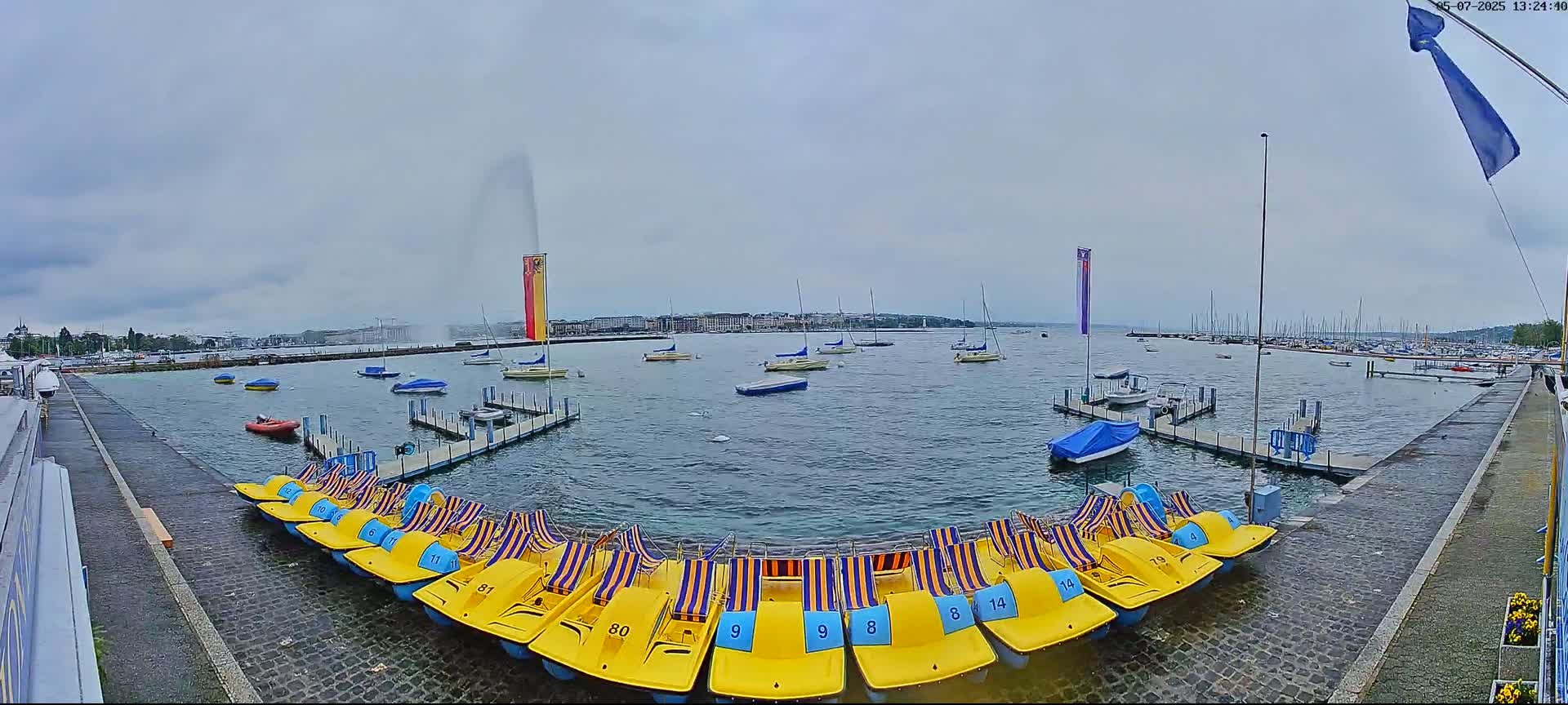 A harbor with numerous small boats and pedal boats, under an overcast sky, features a large water fountain in the distance.
