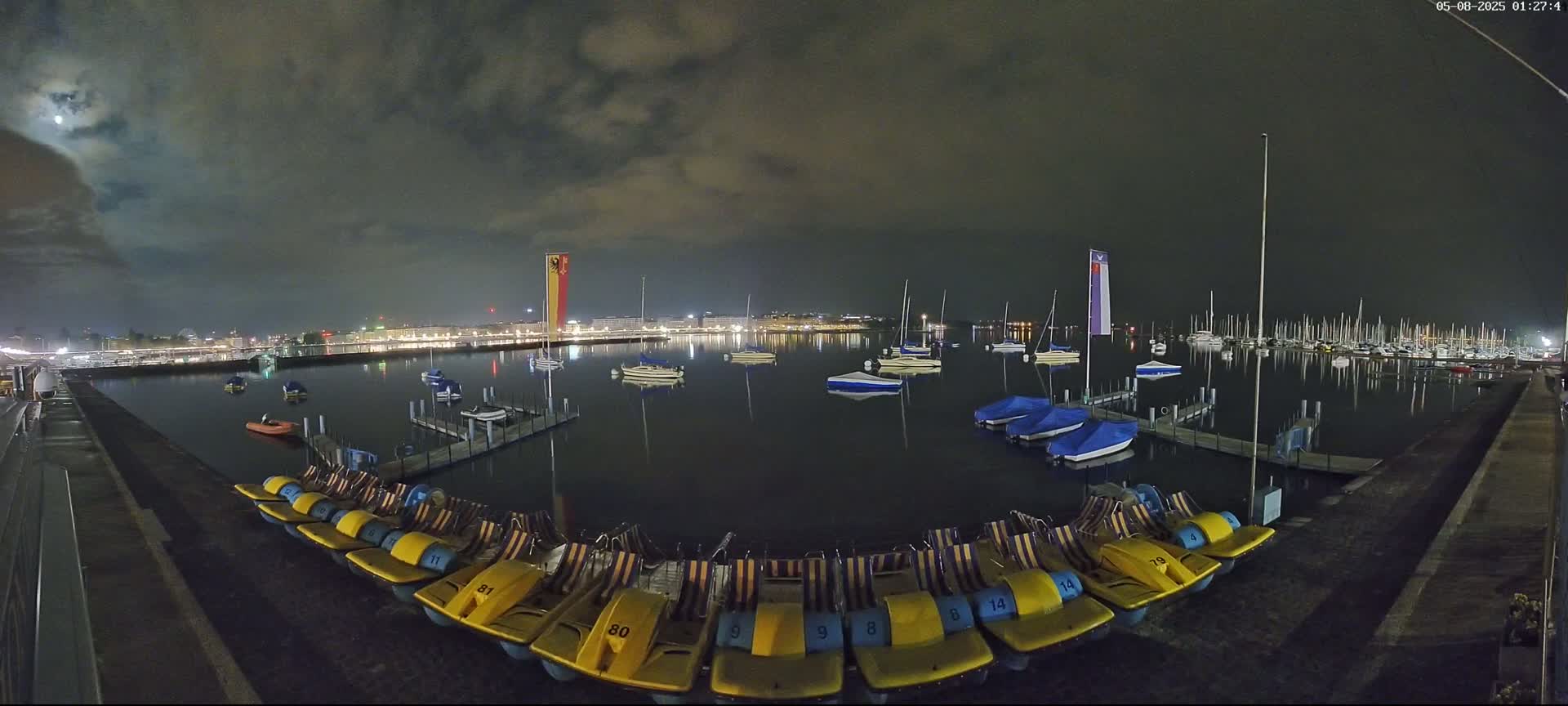 Under a partly cloudy night sky, a calm body of water is filled with docked sailboats and paddle boats, with a city skyline visible in the distance.