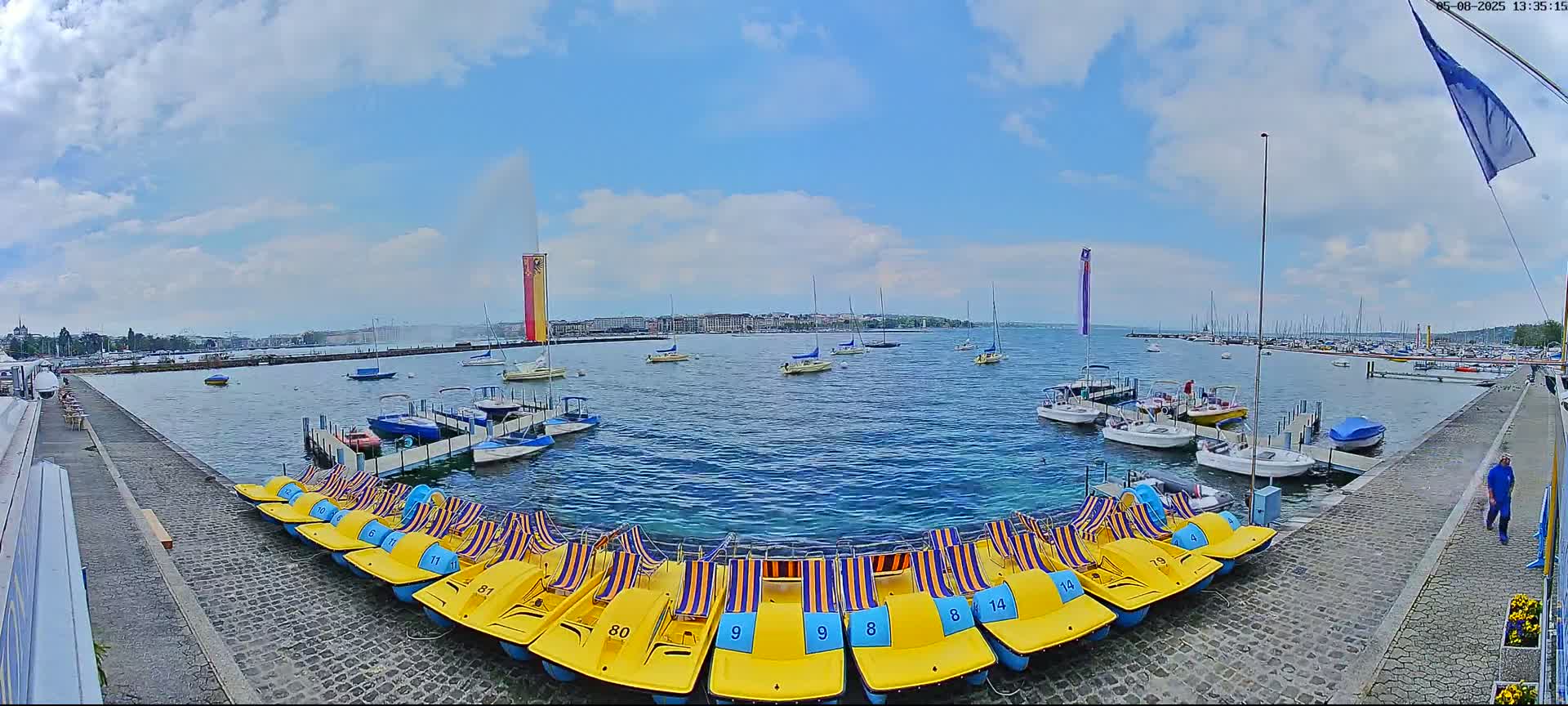 A partly cloudy day shows a harbor with numerous sailboats and pedal boats lined up along a cobblestone walkway, a water fountain in the distance.