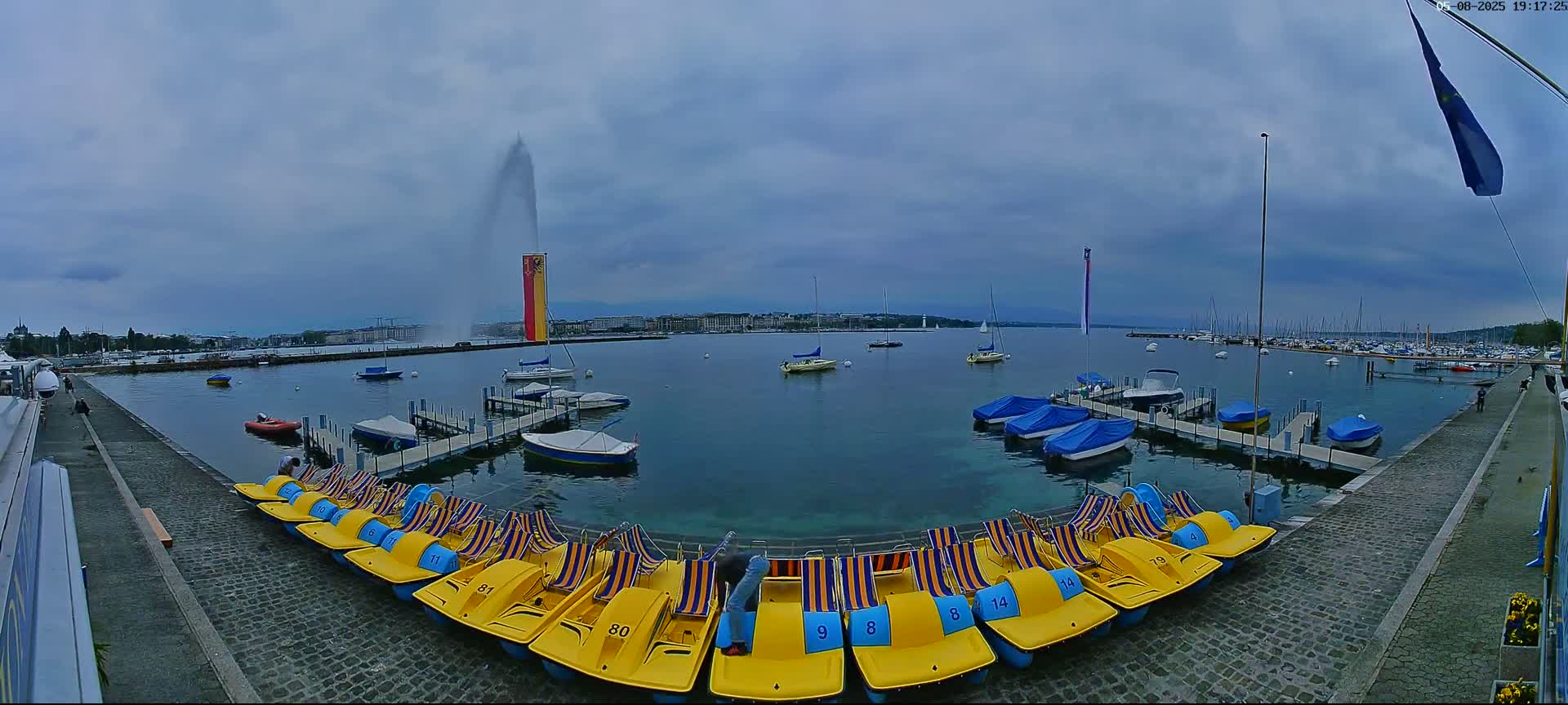 A large water fountain sprays water into the overcast sky above a body of water filled with boats and paddle boats docked along a cobblestone walkway.