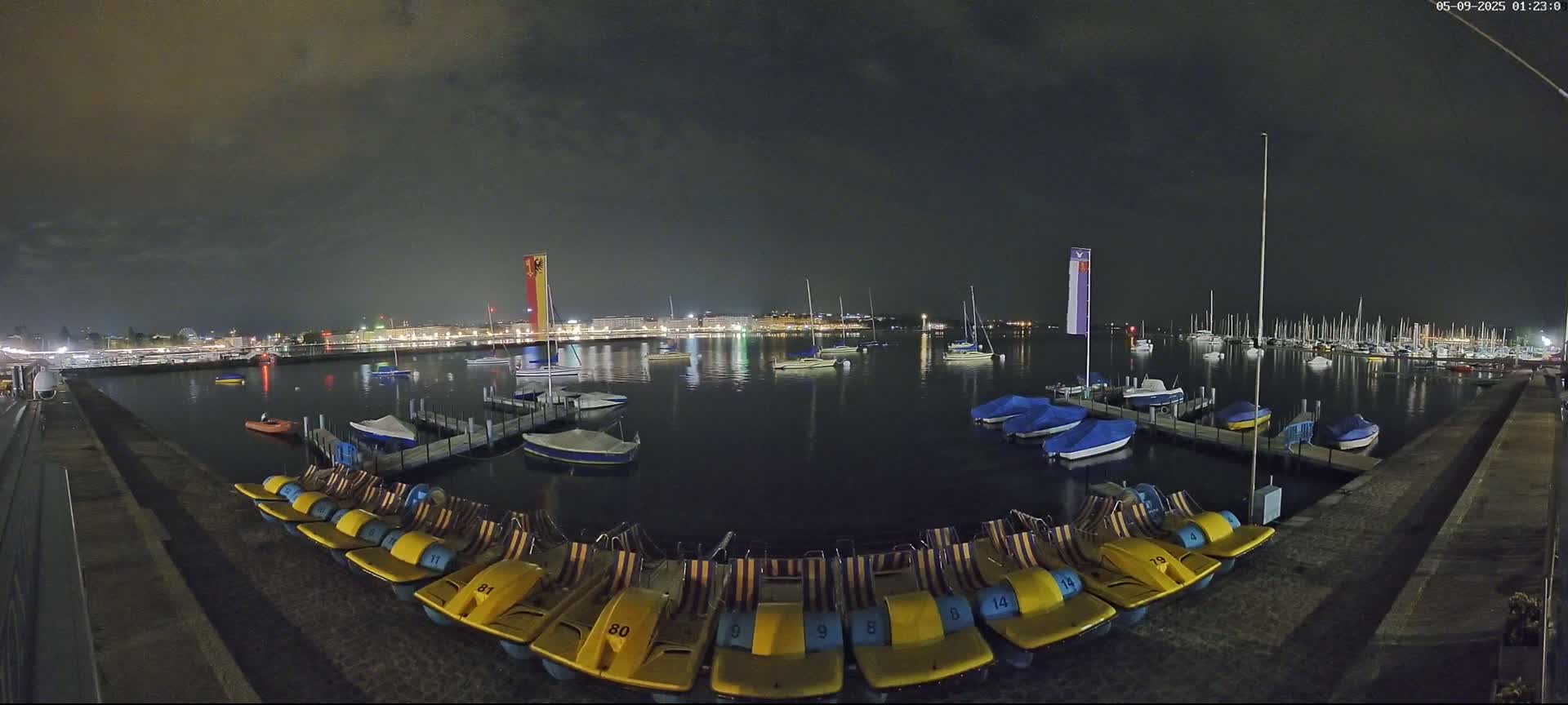 Under a dark, cloudy night sky, a calm body of water shows a marina with numerous sailboats and small motorboats, along with a line of docked pedal boats.