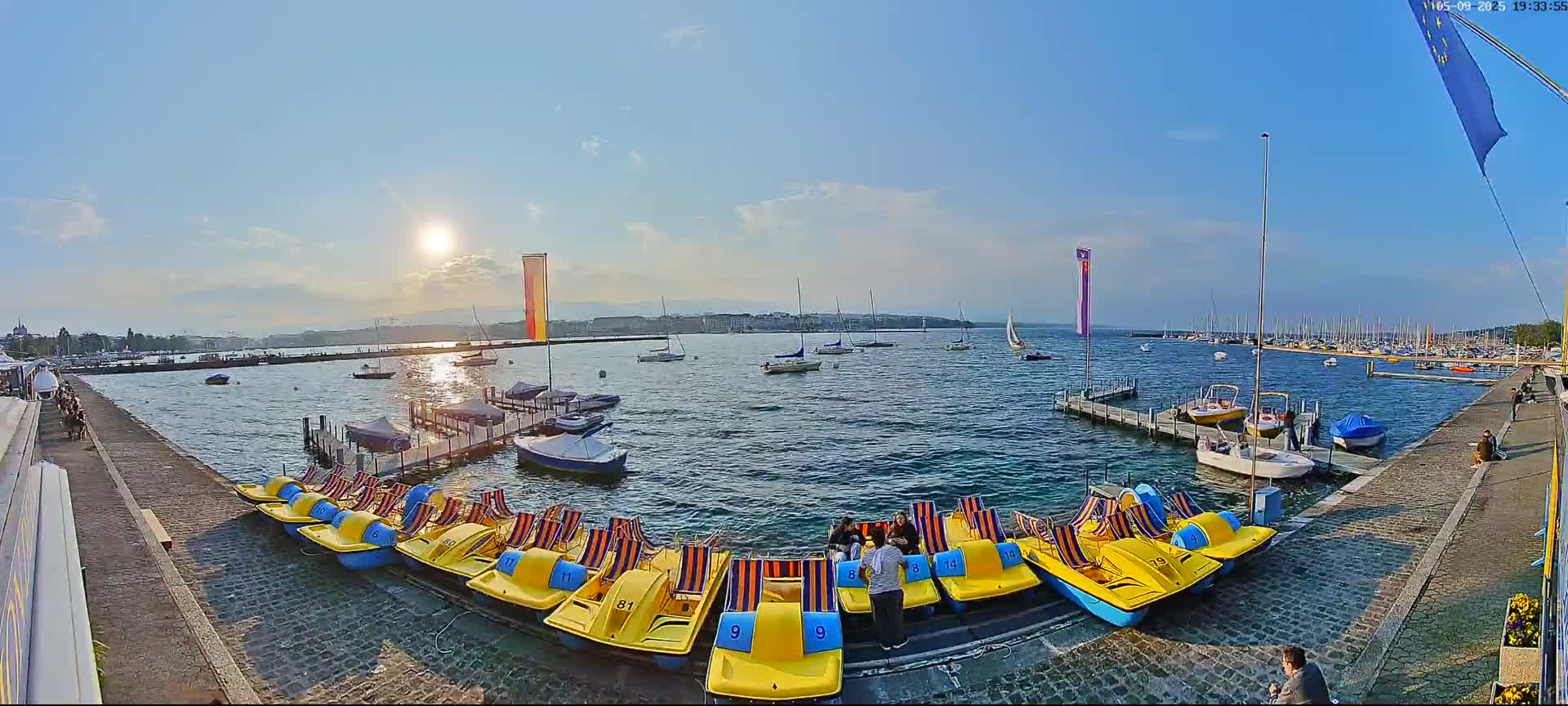 A sunny day on a calm lake shows many boats moored at docks, with several yellow paddle boats lined up along a cobblestone walkway.