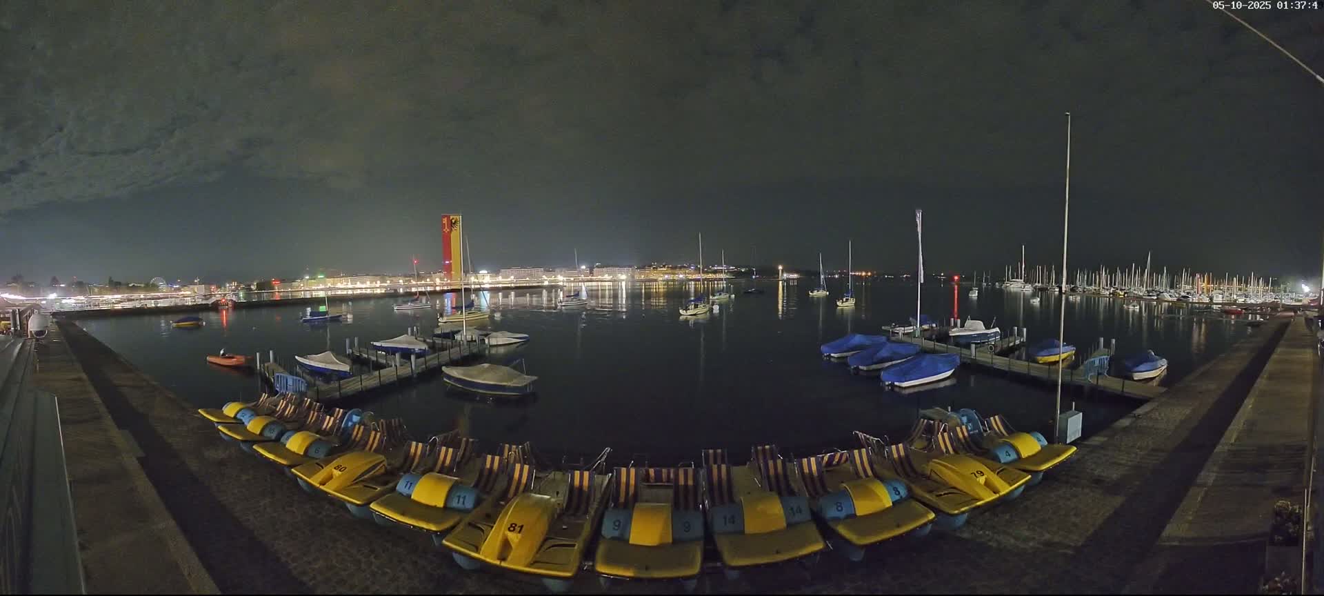 At night, under a mostly cloudy sky, a calm harbor holds numerous docked sailboats and small paddle boats, with a city's lights visible in the distance.