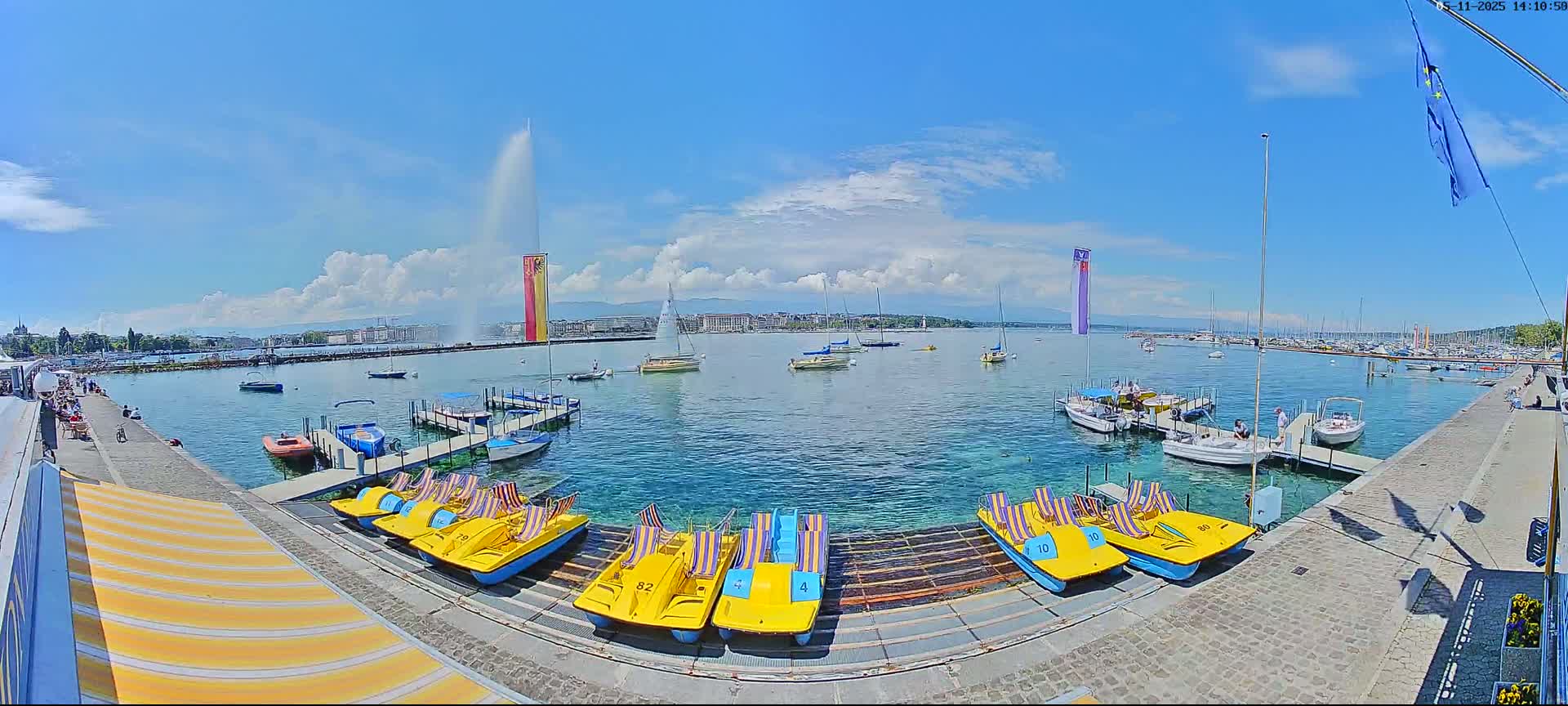 A sunny day on a lake shows a waterfront with several docked boats, pedal boats, and a large fountain in the distance.