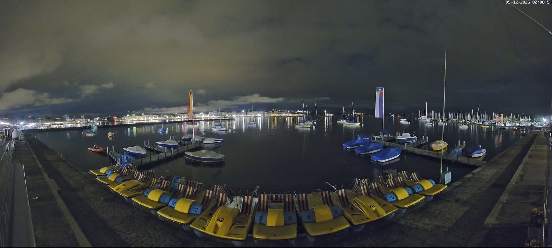 A nighttime view of a harbor with numerous boats and pedal boats docked, under a cloudy sky.