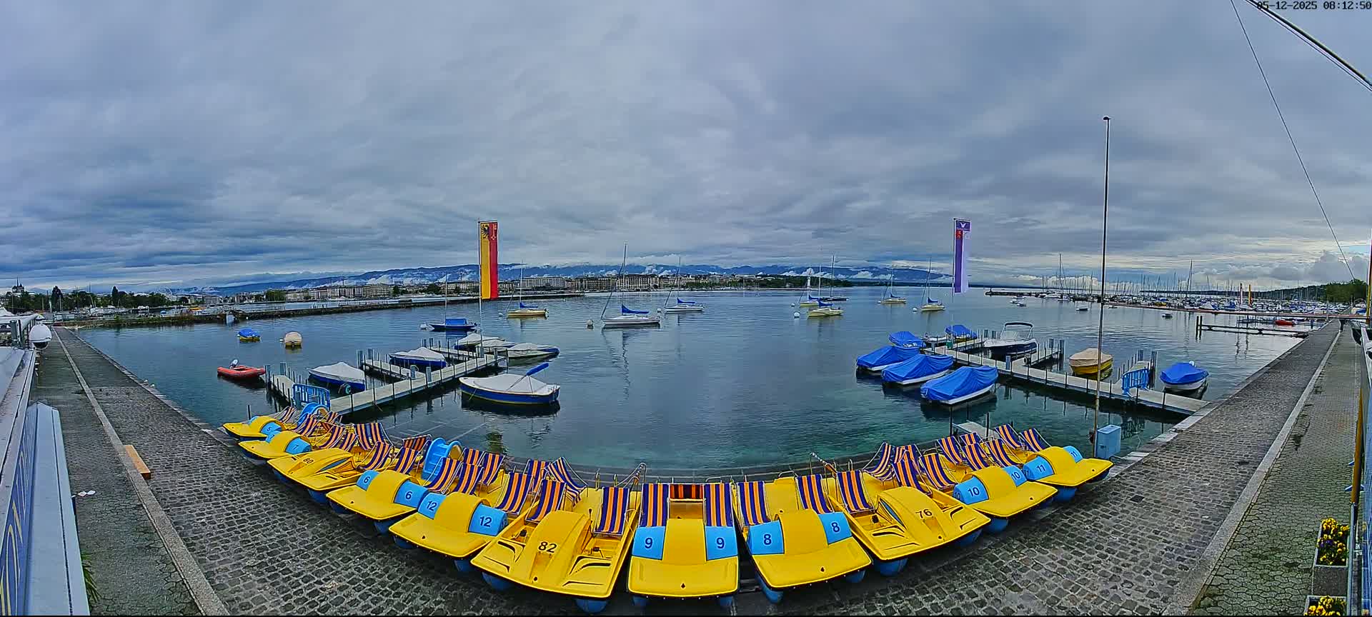 A calm body of water with numerous boats and pedal boats docked along a stone pathway, under a cloudy sky and with mountains in the distance.