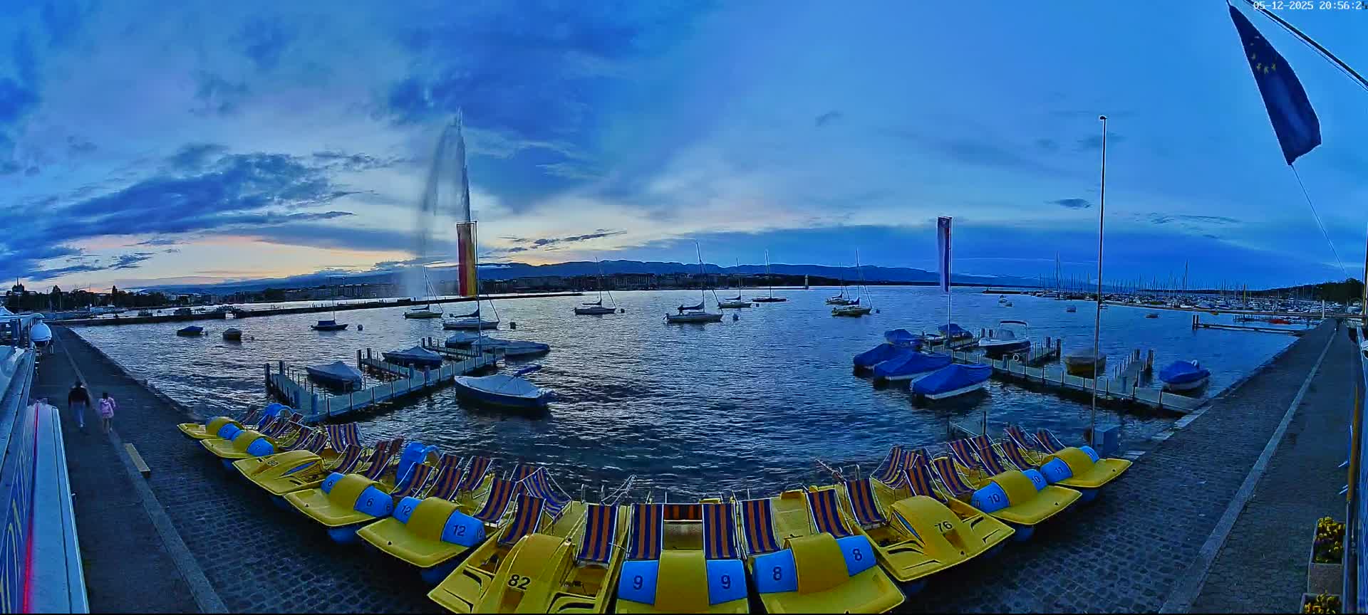 A large water fountain sprays water into the twilight sky over a calm lake filled with docked sailboats and paddle boats.