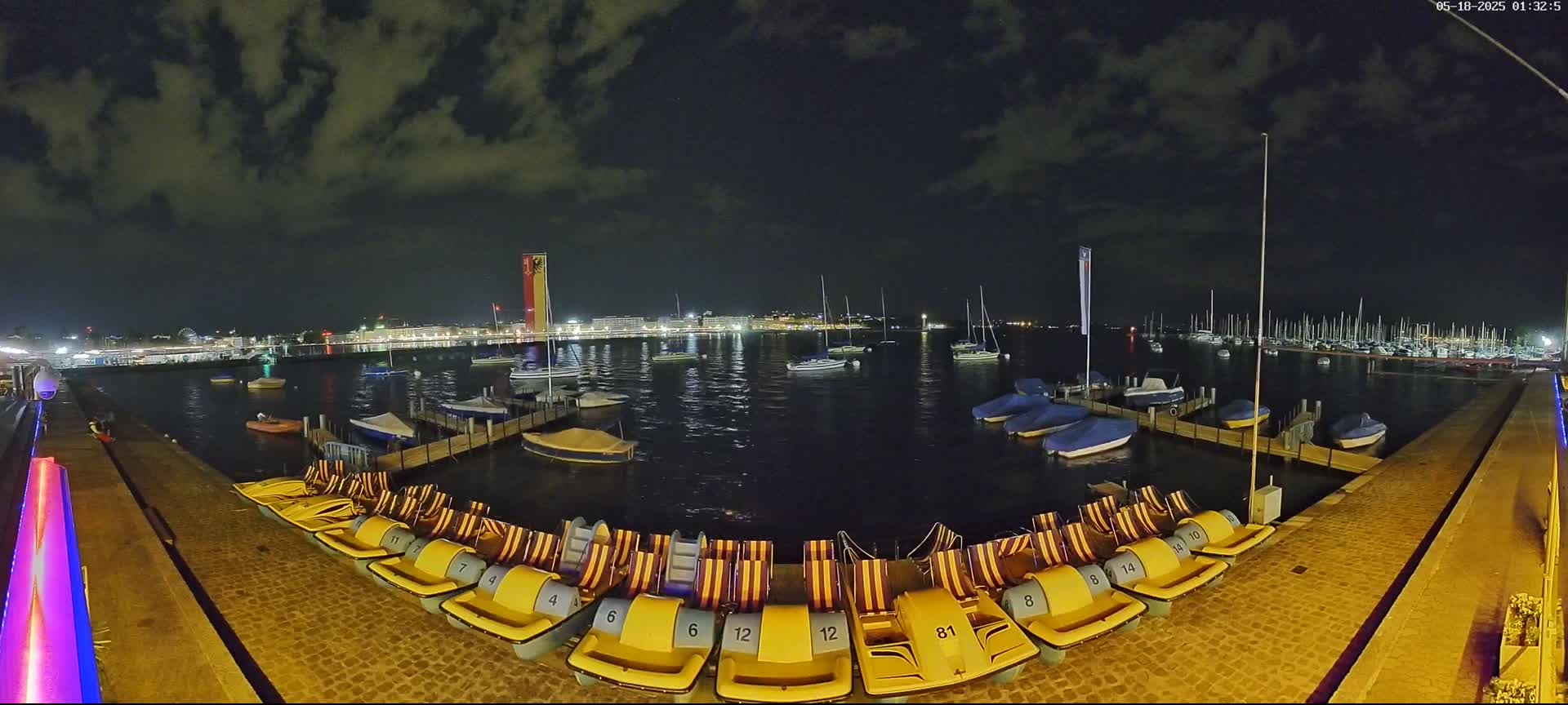 A nighttime view of a harbor with numerous docked boats and paddle boats, under a partly cloudy sky.