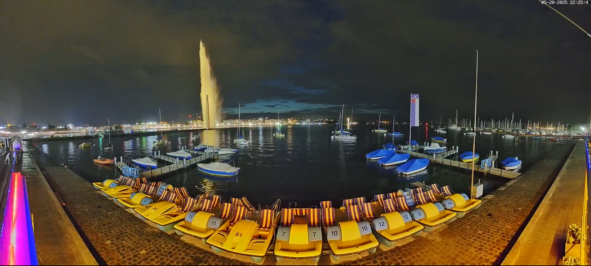At night, a tall fountain sprays water high into the air over a body of water where numerous boats and pedal boats are docked.