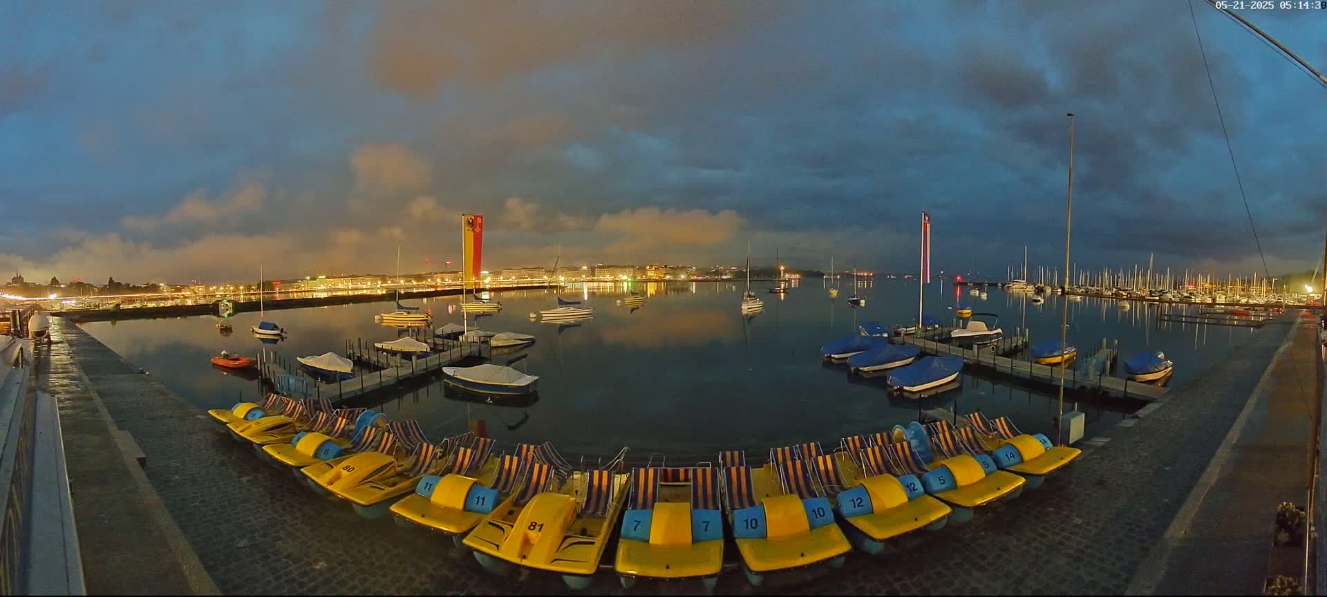 A calm body of water at twilight is dotted with sailboats and pedal boats, with a city skyline illuminated in the background under a partly cloudy sky.