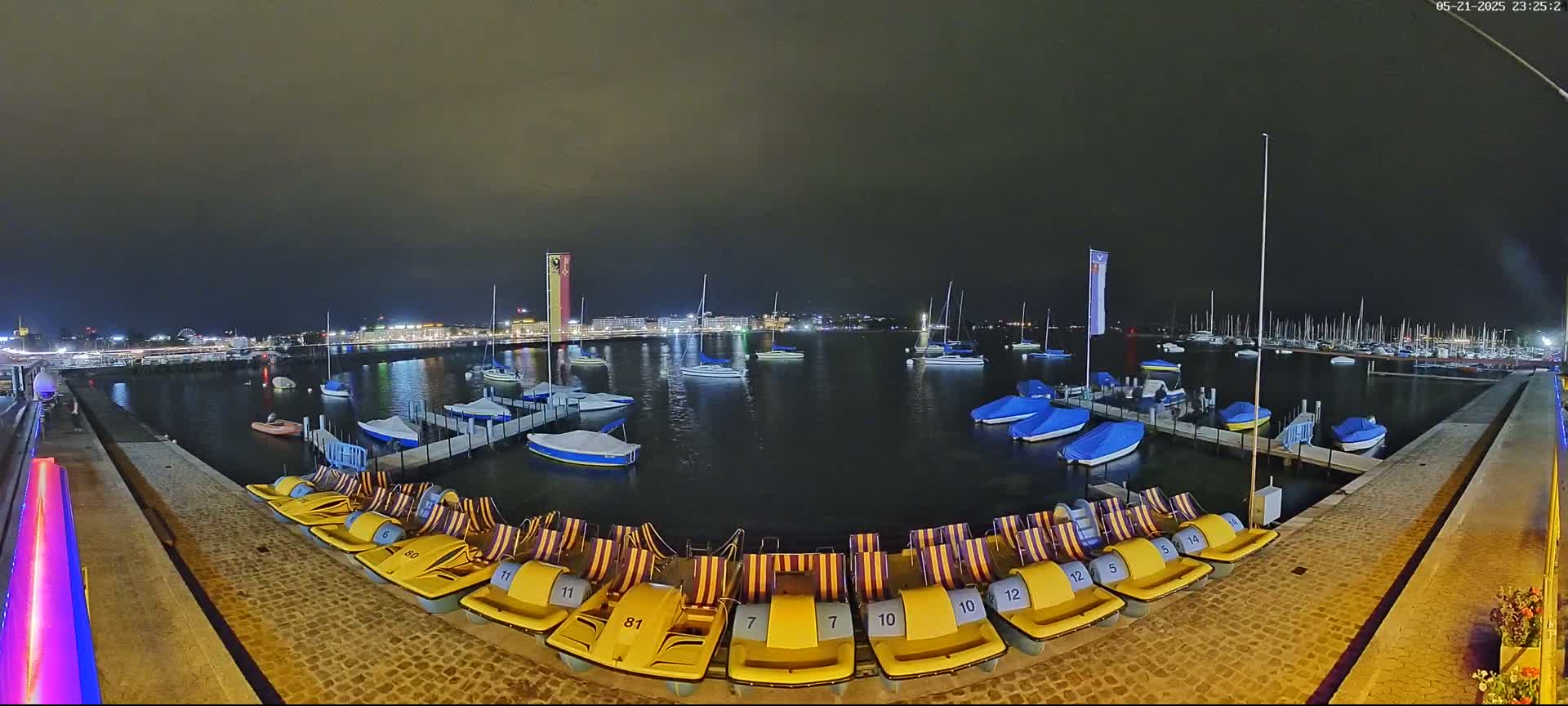 At night, under a dark sky, a harbor filled with sailboats and covered motorboats is shown, with numerous yellow paddle boats lined up on a dock.
