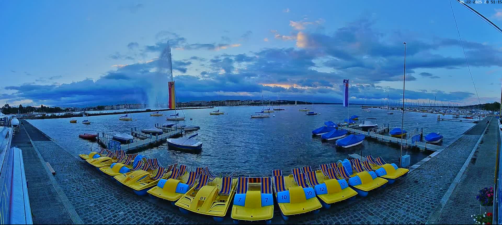 A large water fountain sprays water into the twilight sky over a harbor filled with boats and pedal boats docked at piers.