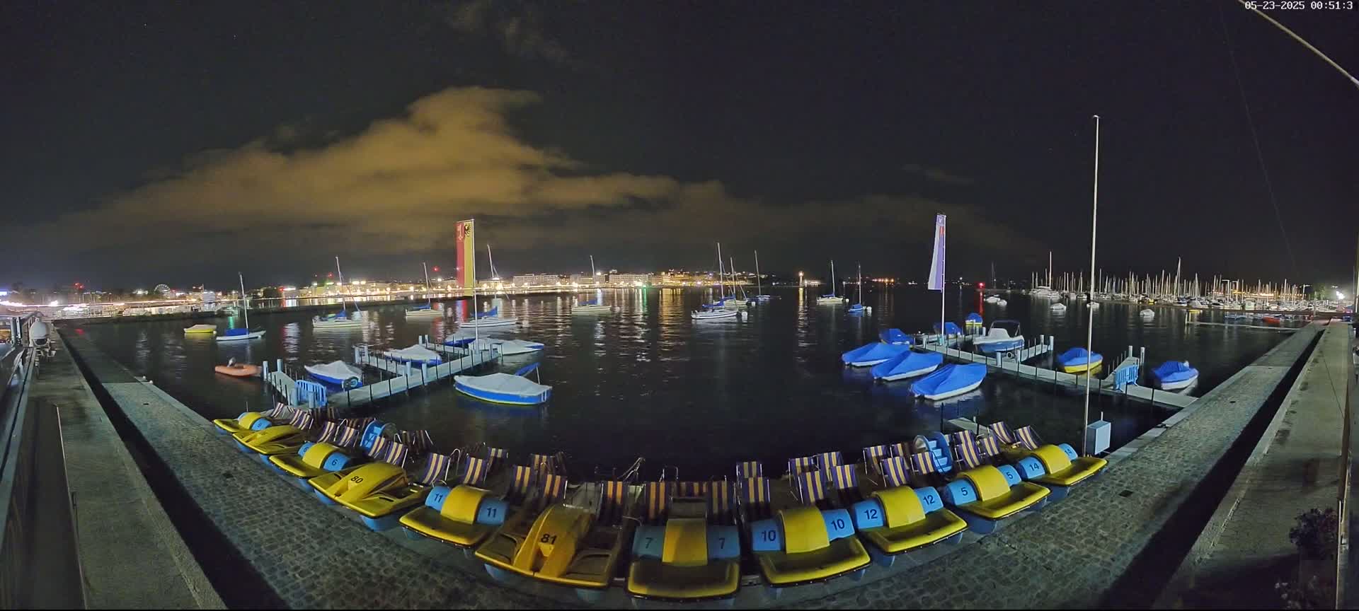 Under a partly cloudy night sky, a calm body of water is filled with moored sailboats and pedal boats docked along a paved waterfront.