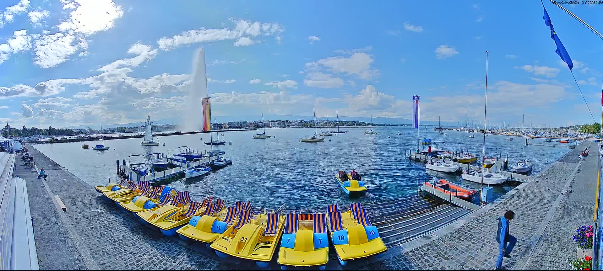 A waterfront scene on a partly sunny day shows numerous boats moored at docks, several paddle boats on the shore, and a large fountain spraying water into the air in the background.