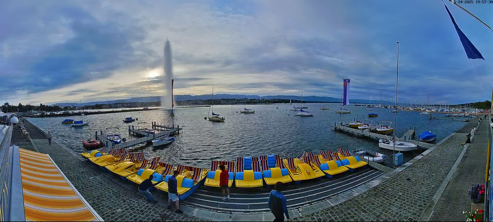 A tall fountain sprays water into the partly cloudy sky above a calm lake with numerous docked boats and pedal boats on the shore.