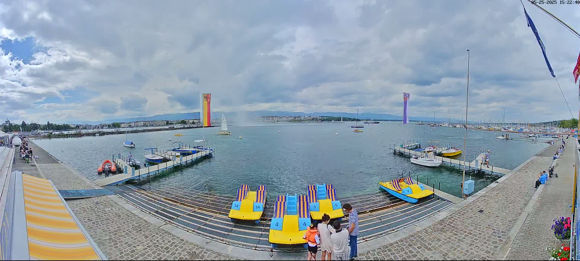 A partly cloudy day shows a lake harbor with several small boats and pedal boats at docks, a water fountain in the distance, and people near the water's edge.