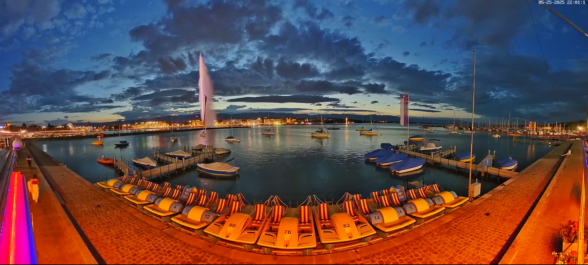 Under a partly cloudy twilight sky, a large water fountain sprays water high into the air over a calm lake harbor filled with boats and docked pedal boats.